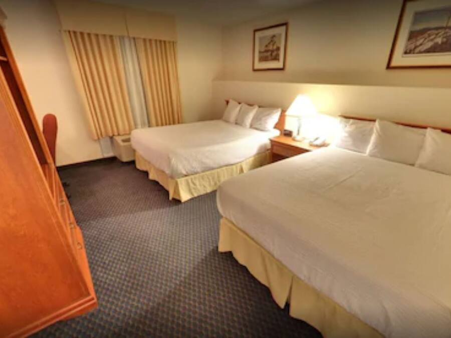 A hotel room with two beds, a wooden wardrobe, a nightstand with a lamp, and a window with curtains.