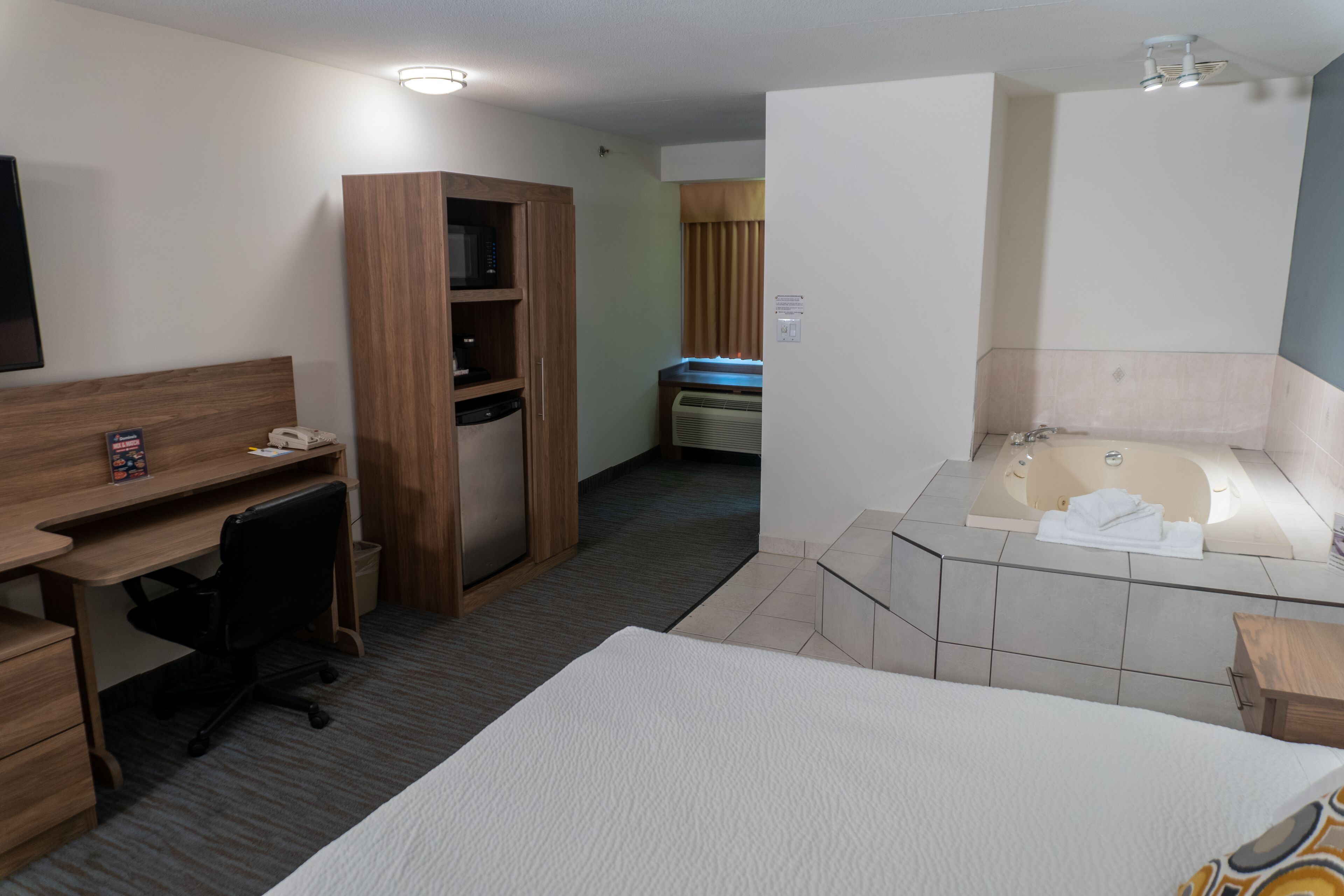 A hotel room with a bed, a desk with a computer, a wardrobe, a bathroom with a bathtub, and a window with curtains.
