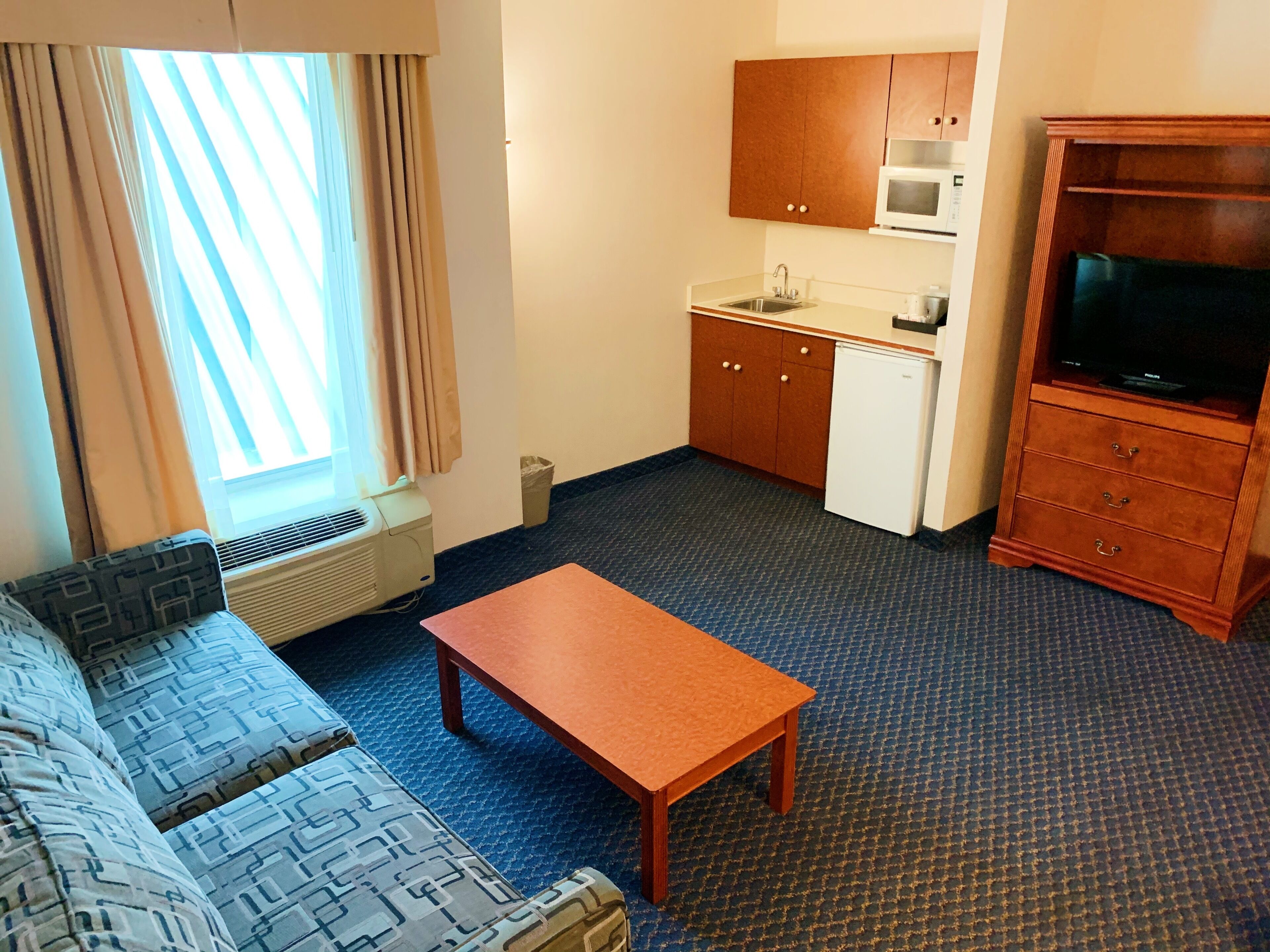 A compact hotel room with a sofa, a wooden table, a kitchenette, and a television.