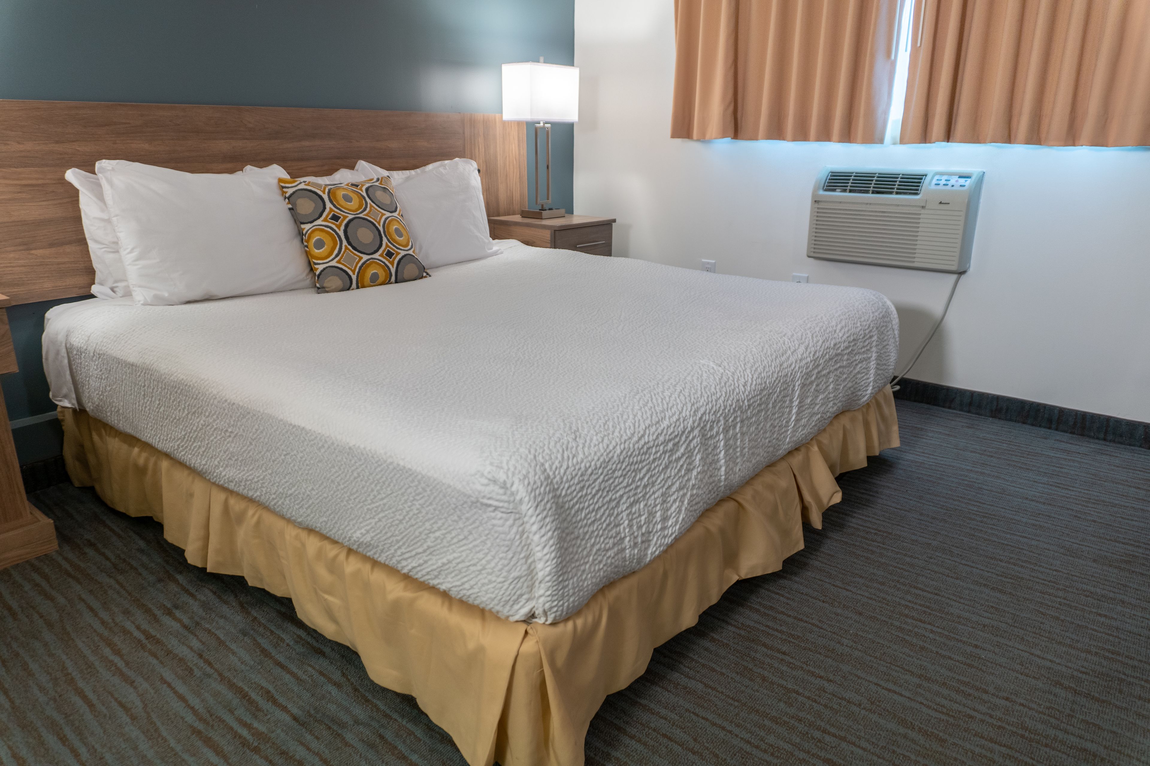 A neatly made bed with a white comforter and a decorative pillow, a bedside lamp, and a wall-mounted air conditioner.