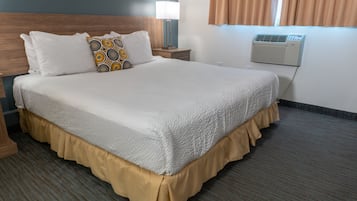 A neatly made bed with a white comforter and a decorative pillow, a bedside lamp, and a wall-mounted air conditioner.