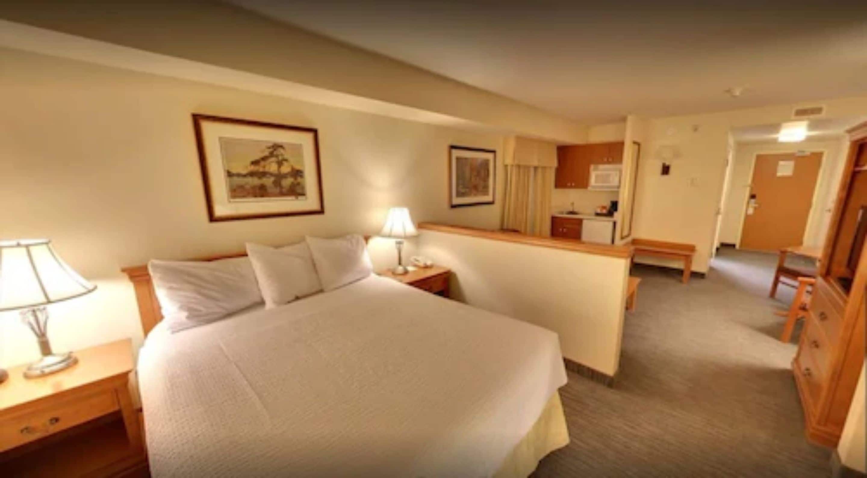 Una habitación de hotel con una cama grande, mesitas de noche con lámparas, un escritorio pequeño con lámpara, una kitchenette y un baño visible a través de una puerta abierta.