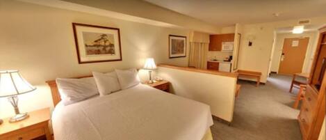 A hotel room with a large bed, bedside tables with lamps, a small desk with a lamp, a kitchenette, and a bathroom visible through an open door.