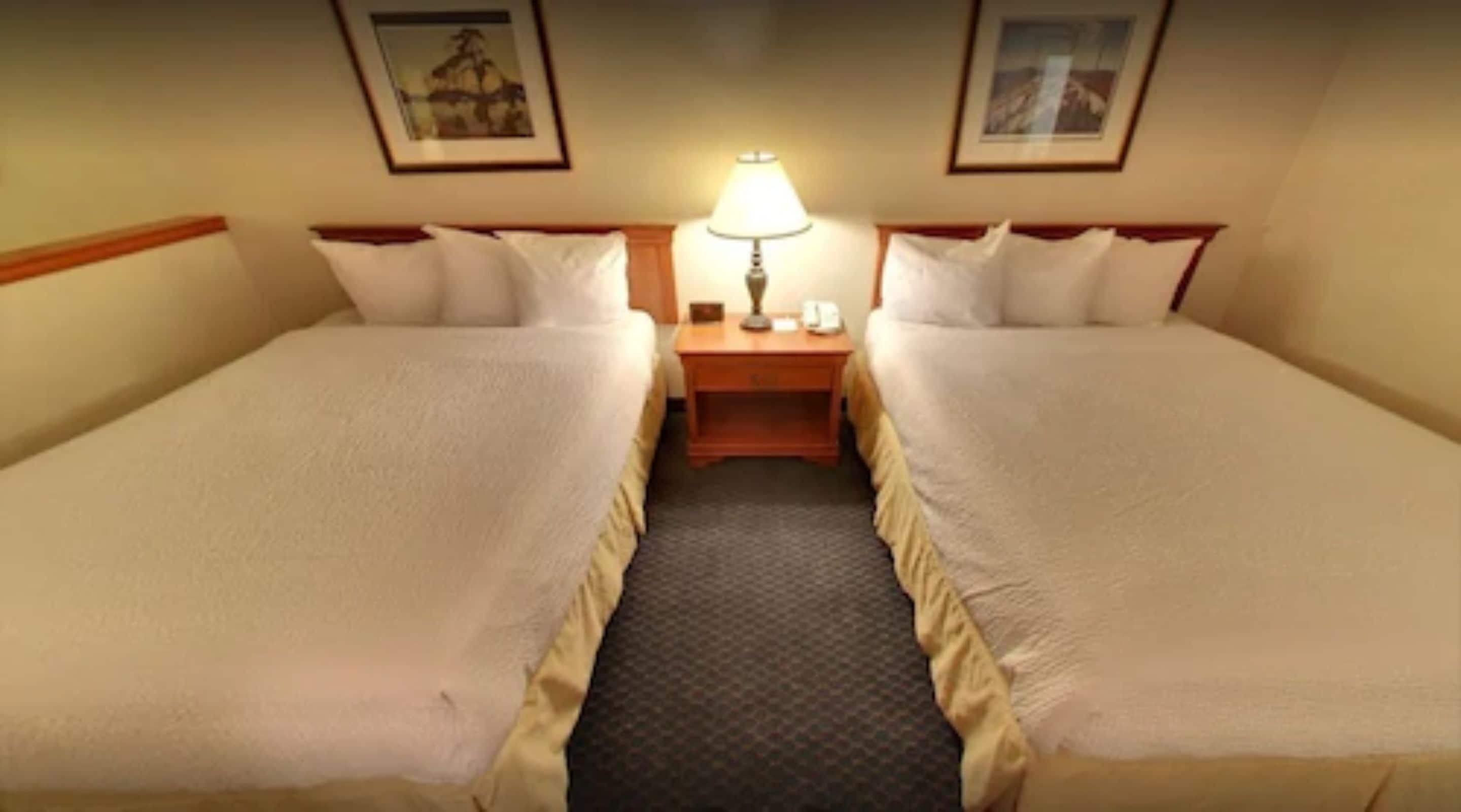 A standard hotel room with two single beds, a nightstand with a lamp, and framed pictures on the walls.
