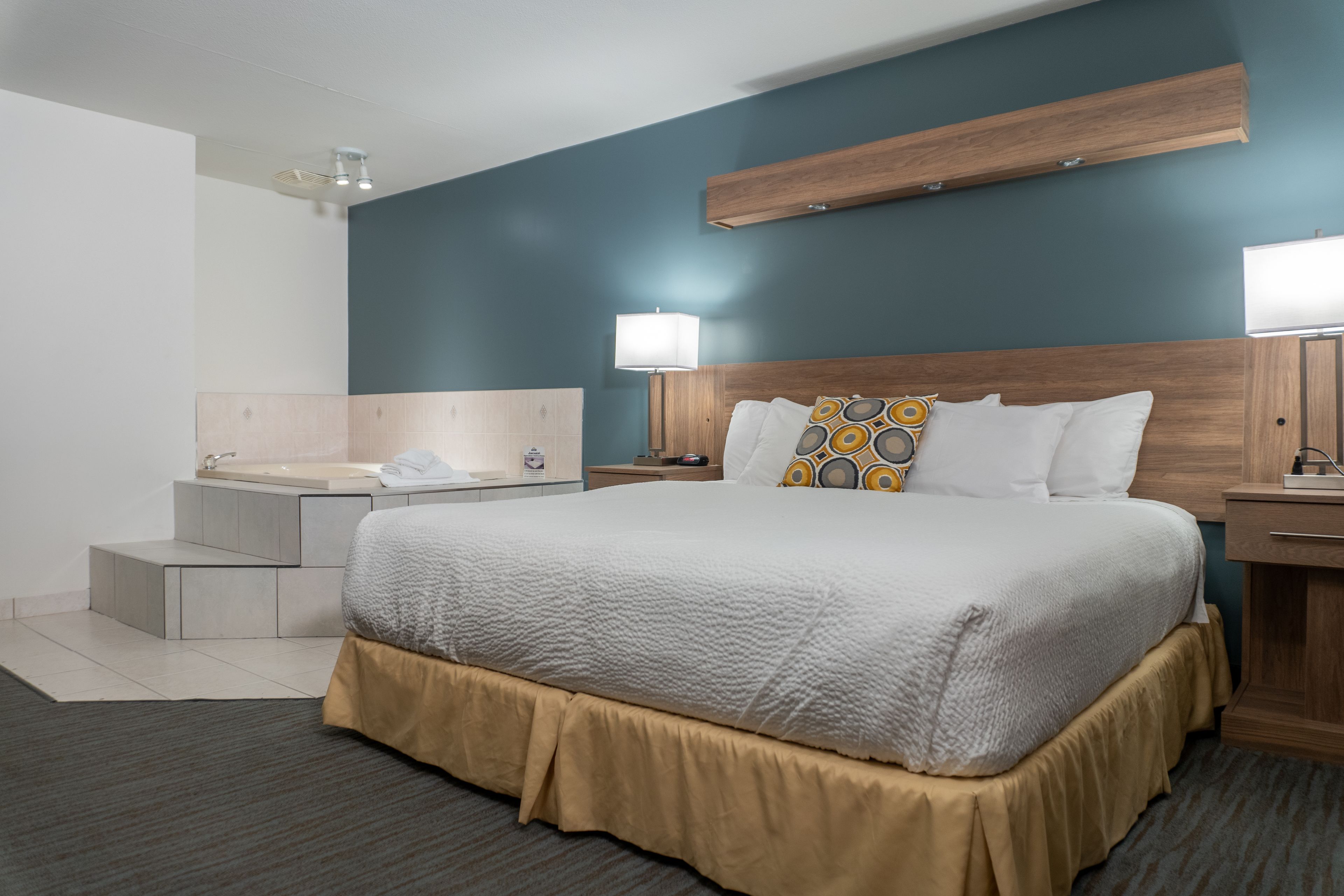 A hotel room with a large bed, a wooden headboard, a bedside table with a lamp, a nightstand, and a built-in shelf above the bed.