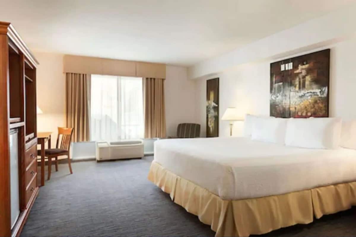 A hotel room with a large bed, a desk, a chair, a window with curtains, and a painting on the wall.