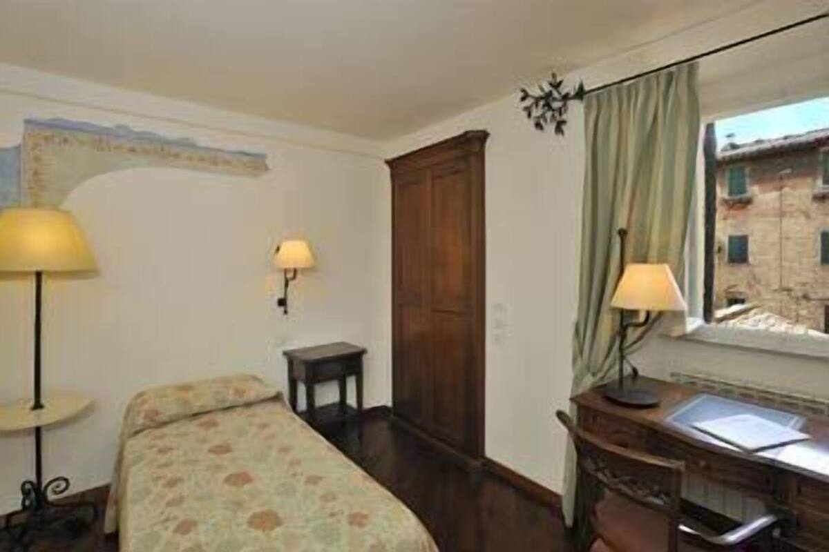 A hotel room with a bed, a desk with a chair, a window with curtains, and a lamp on the desk.