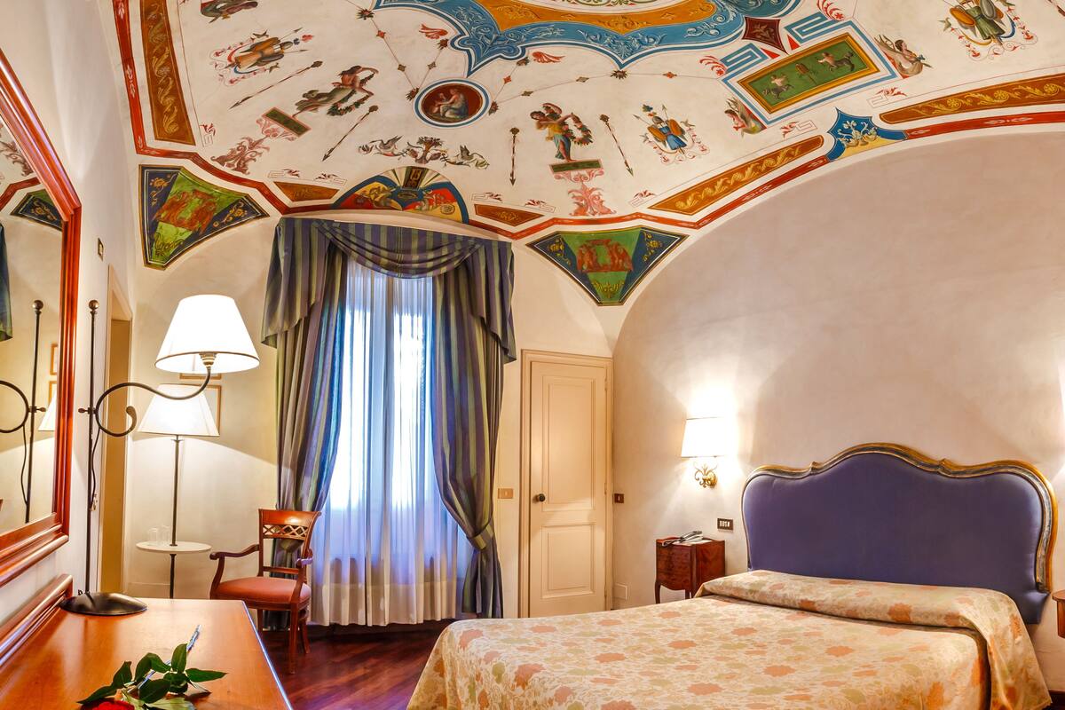 A hotel room with a large bed, a desk with a lamp, a chair, and a window with curtains.