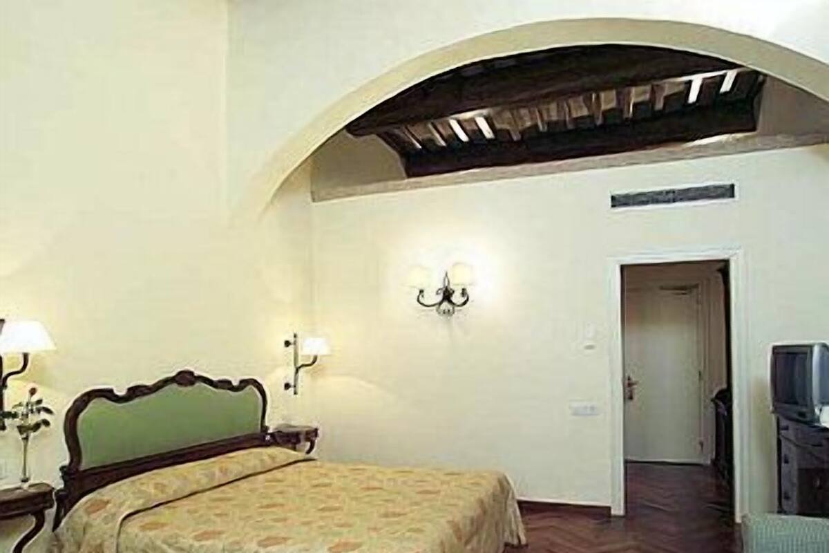 A bedroom with a bed, a nightstand, a lamp, a wall-mounted light fixture, and a television.