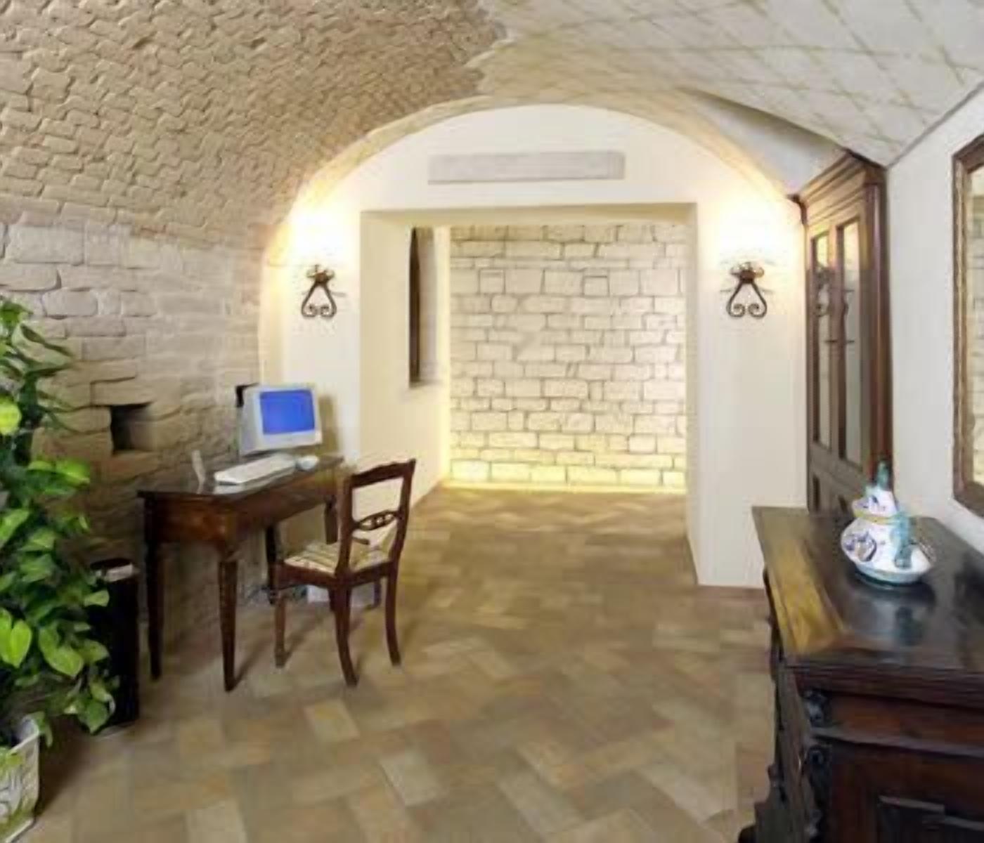 A room with a stone wall, a desk with a computer, a chair, and a small table with a decorative item.