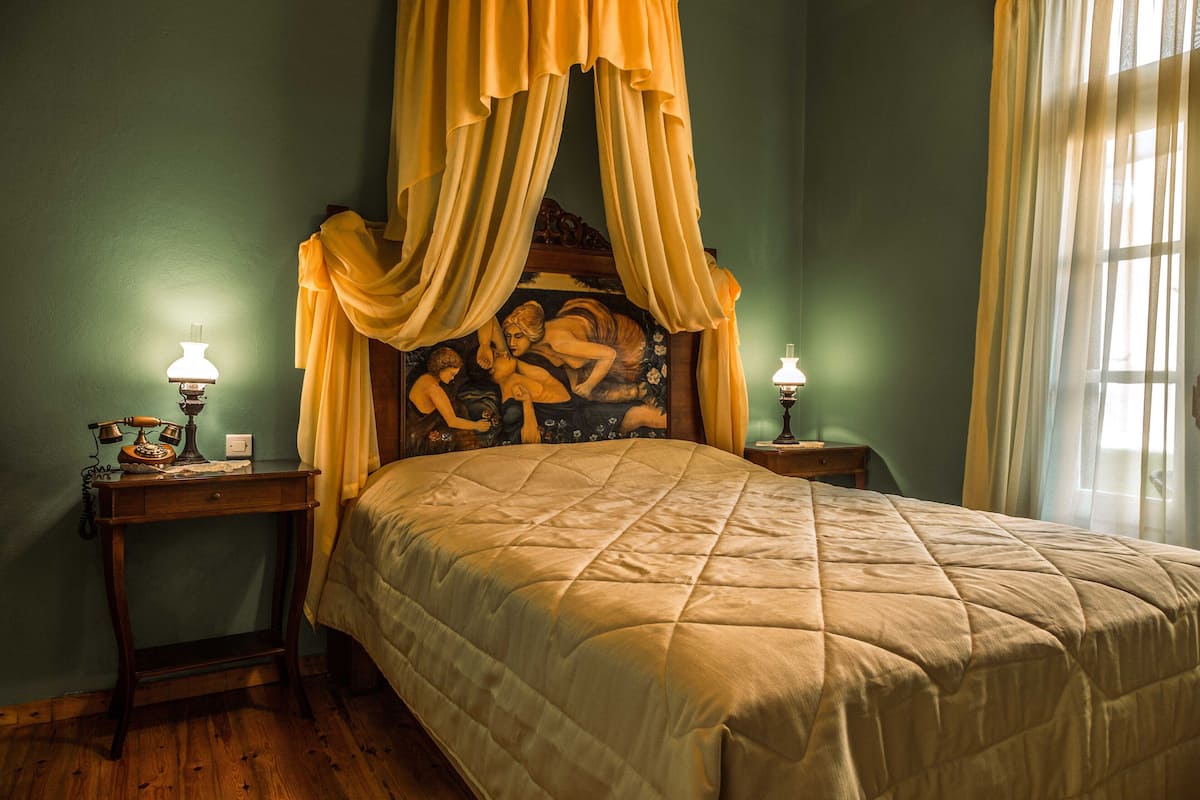 Un dormitorio con cama con dosel, dos mesitas de noche con lámparas y un cuadro en la cabecera.