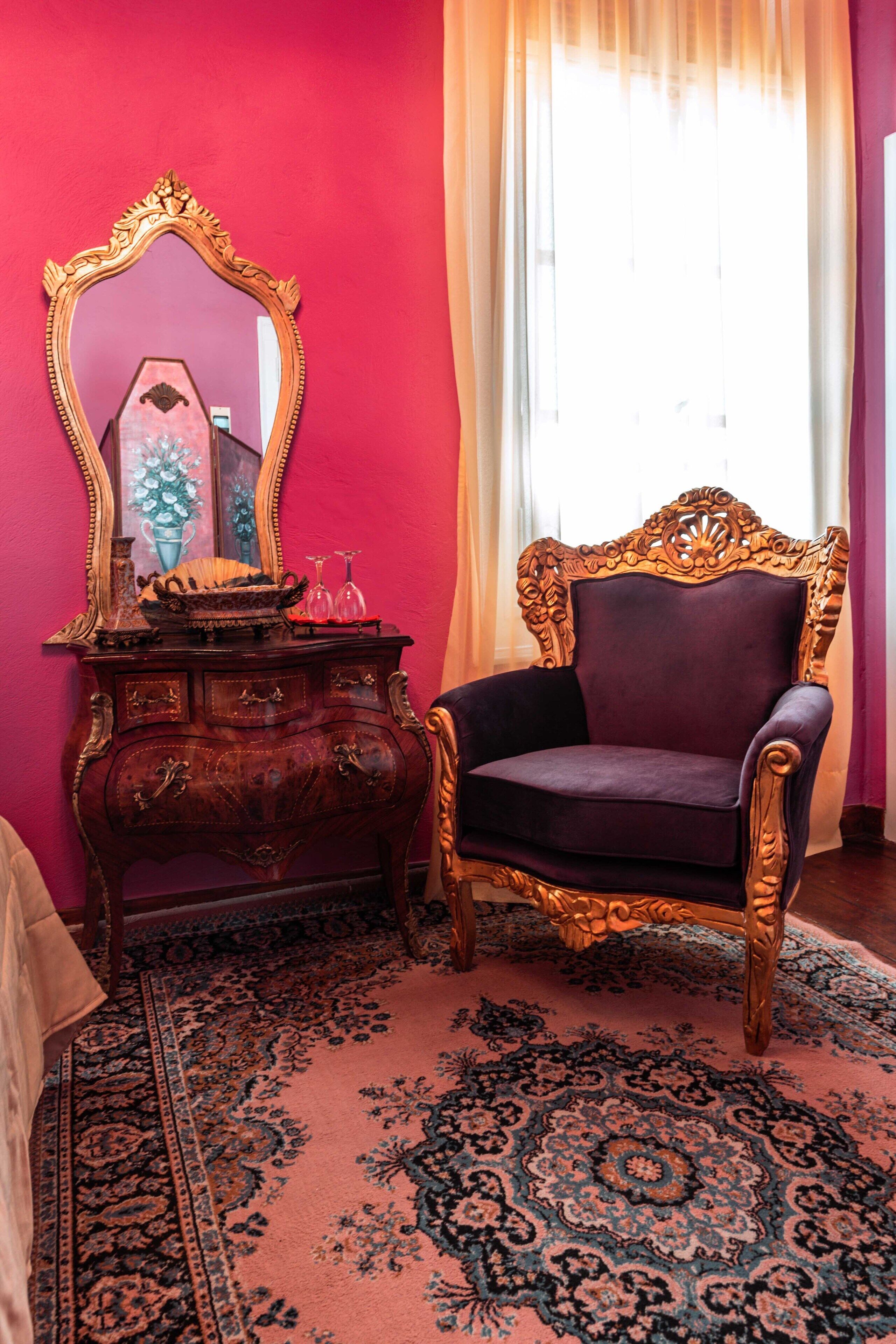 A room with a large ornate mirror, a dark upholstered chair, a wooden dresser with drawers, and a patterned rug.