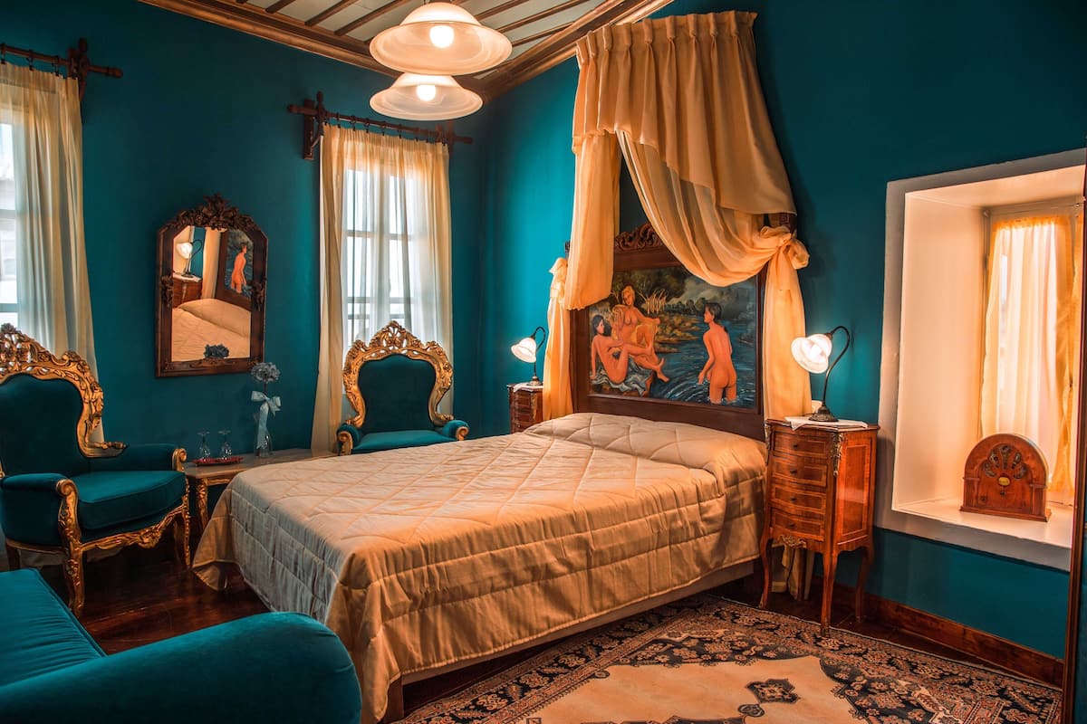 Un dormitorio con una cama grande, dos sillas, una mesita de noche, un aparador, un espejo y una ventana con cortinas.