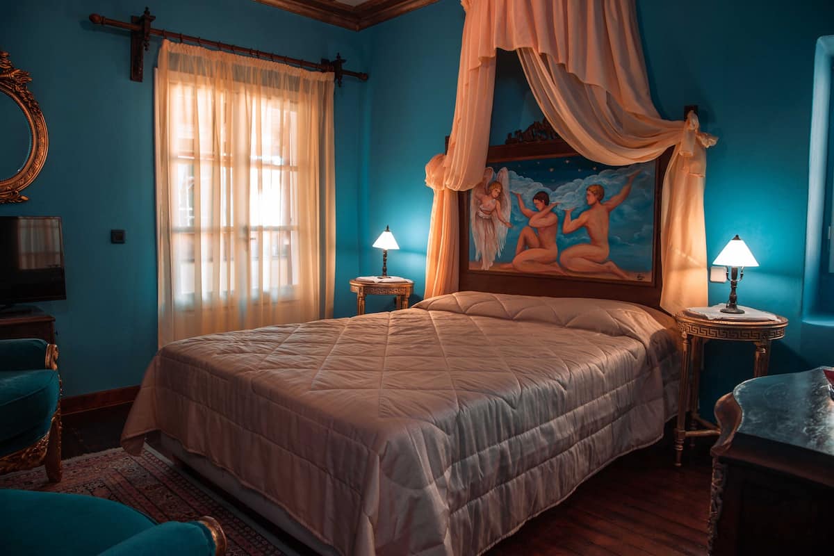 Un dormitorio con una cama grande, dos mesitas de noche con lámparas, un cuadro de querubines, un espejo y una ventana con cortinas traslúcidas.