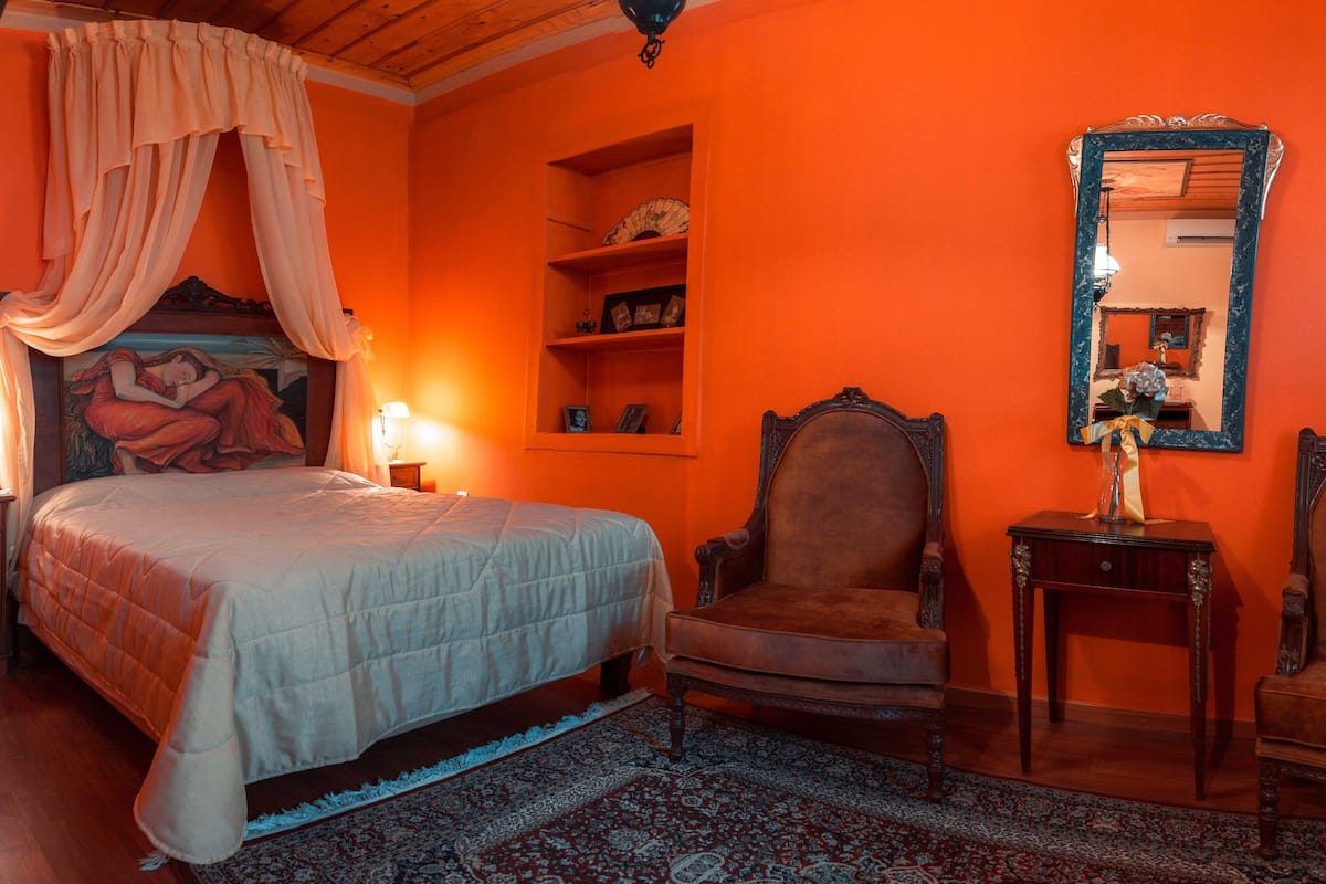 Un dormitorio con una cama grande, una silla, una mesita de noche, un espejo y un estante con objetos decorativos.