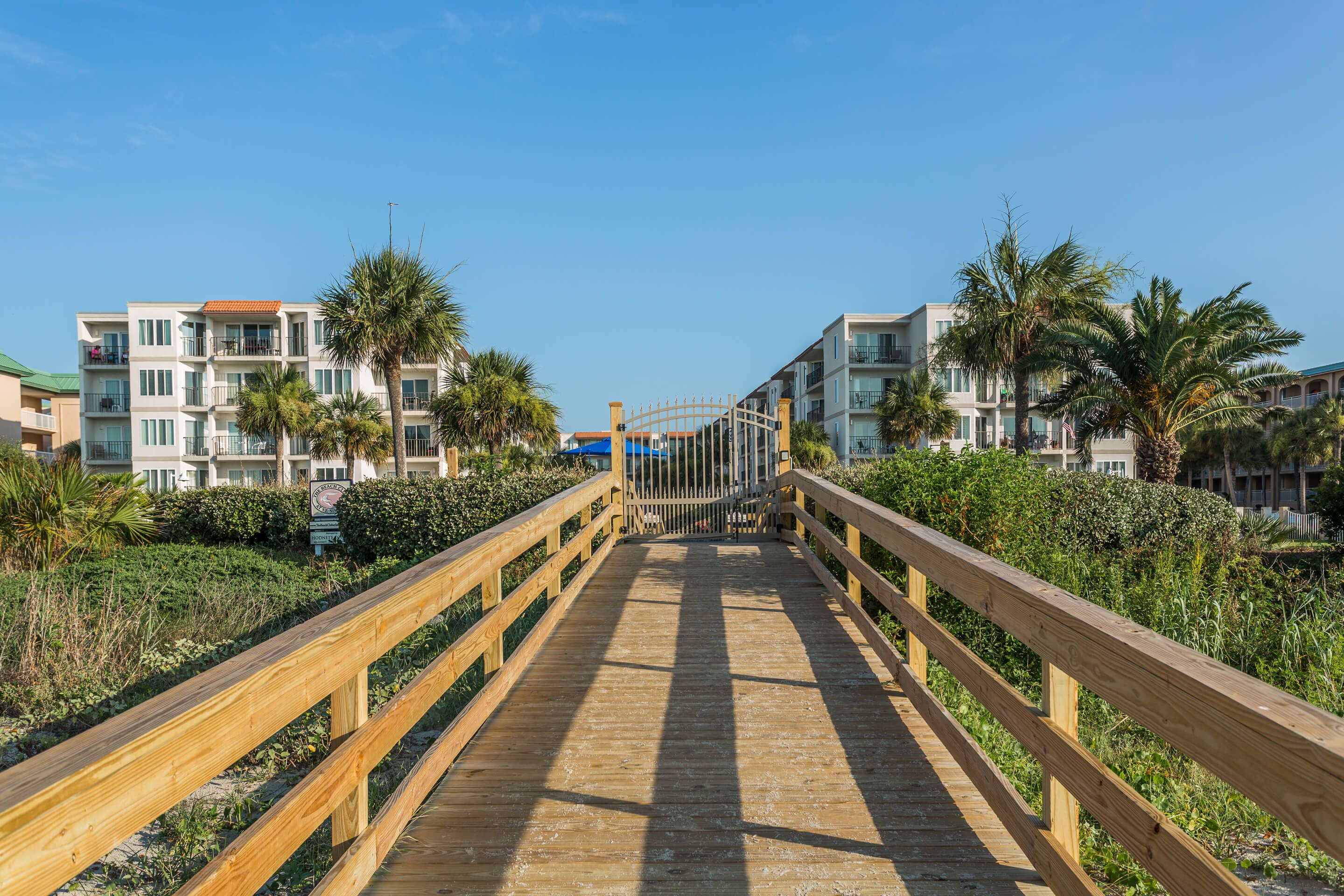 A wooden boardwalk leading to a gated entrance with a view of multi-story buildings and palm trees.