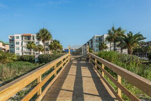 A wooden boardwalk leading to a gated entrance with a view of multi-story buildings and palm trees. - Hodnett Cooper's Beach Club (St. Simons Island)