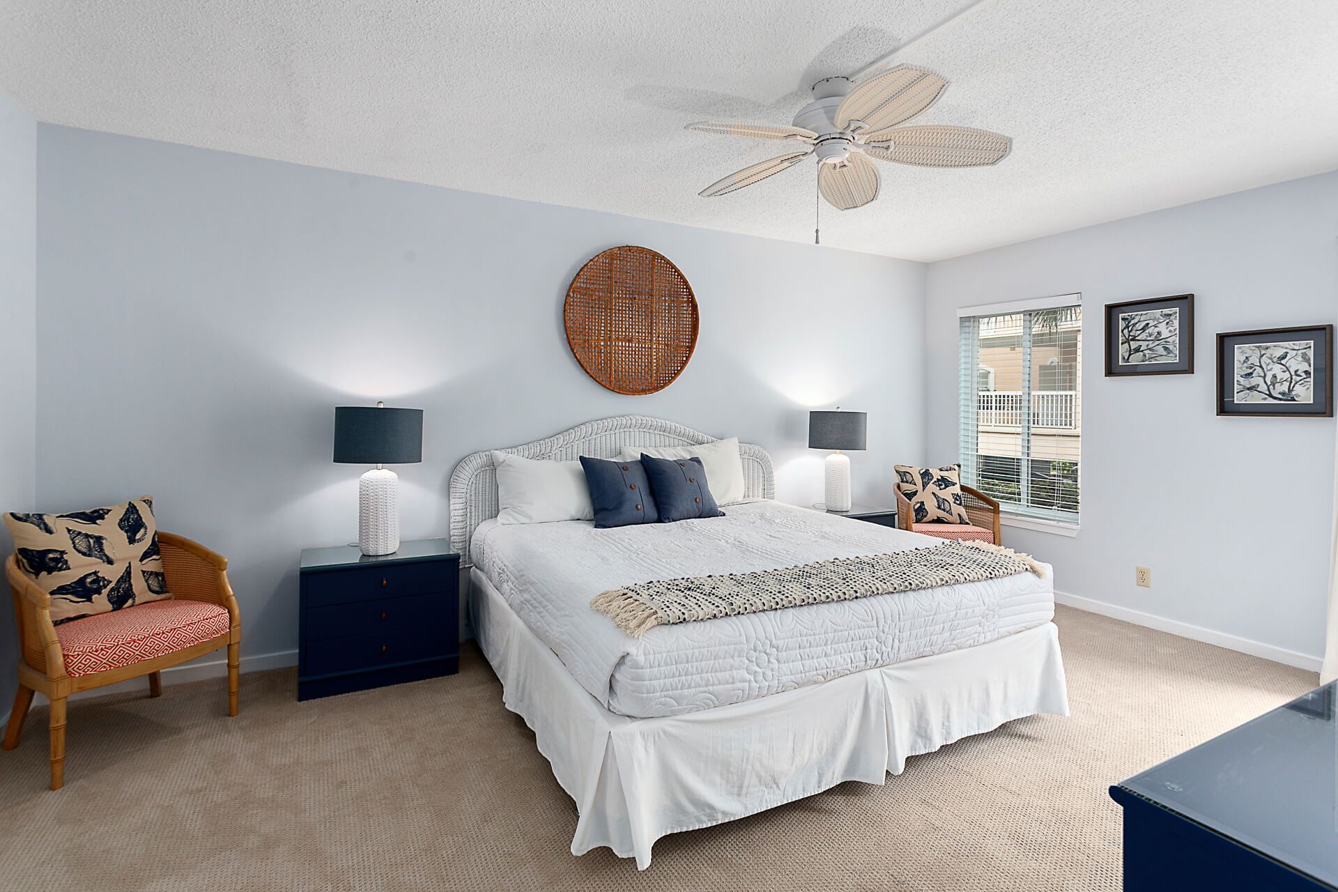 A bedroom with a large bed, two bedside tables with lamps, a rattan chair, a ceiling fan, and a window with blinds.