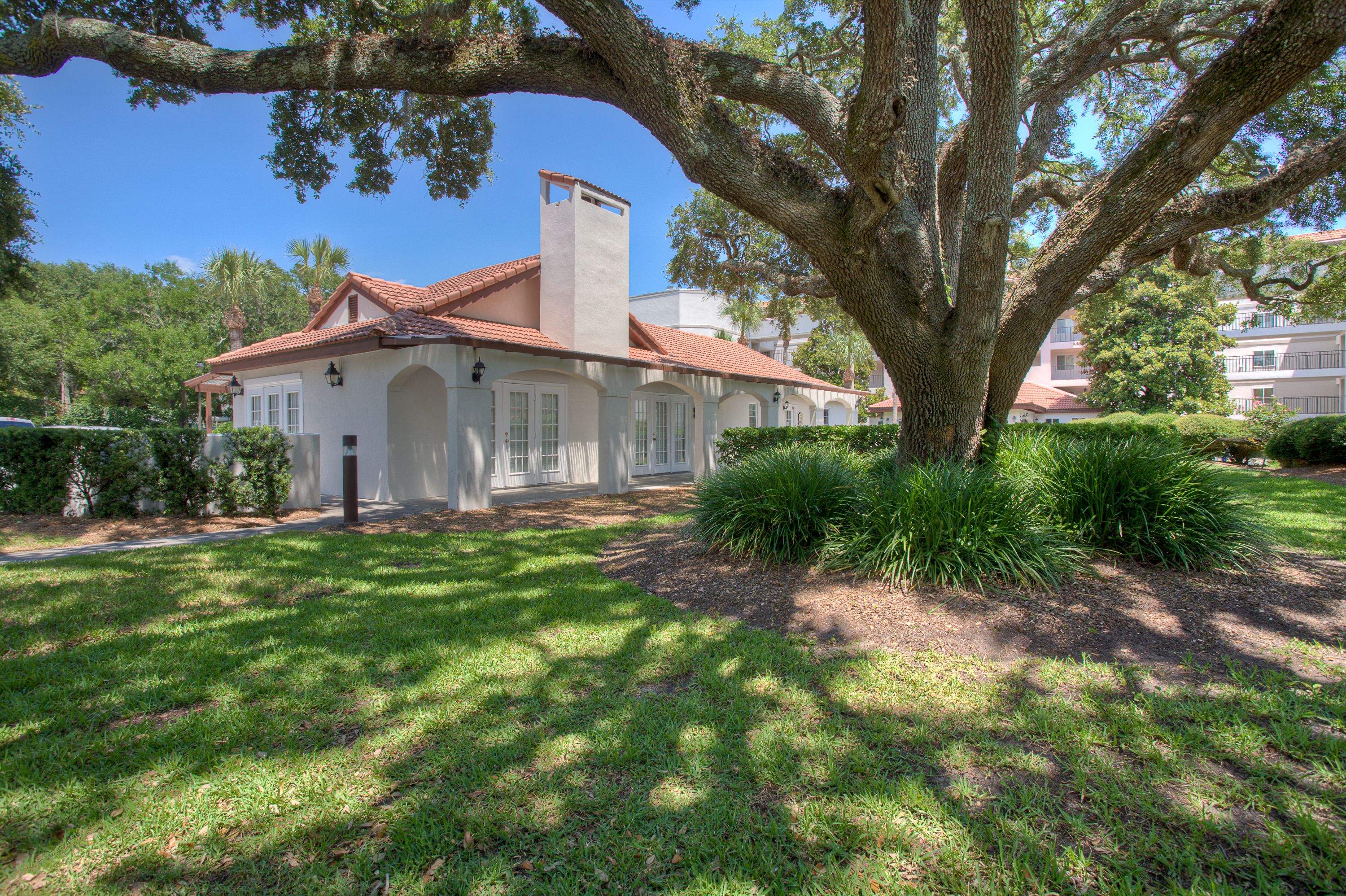 A white building with a red-tiled roof, surrounded by lush greenery and a large tree.