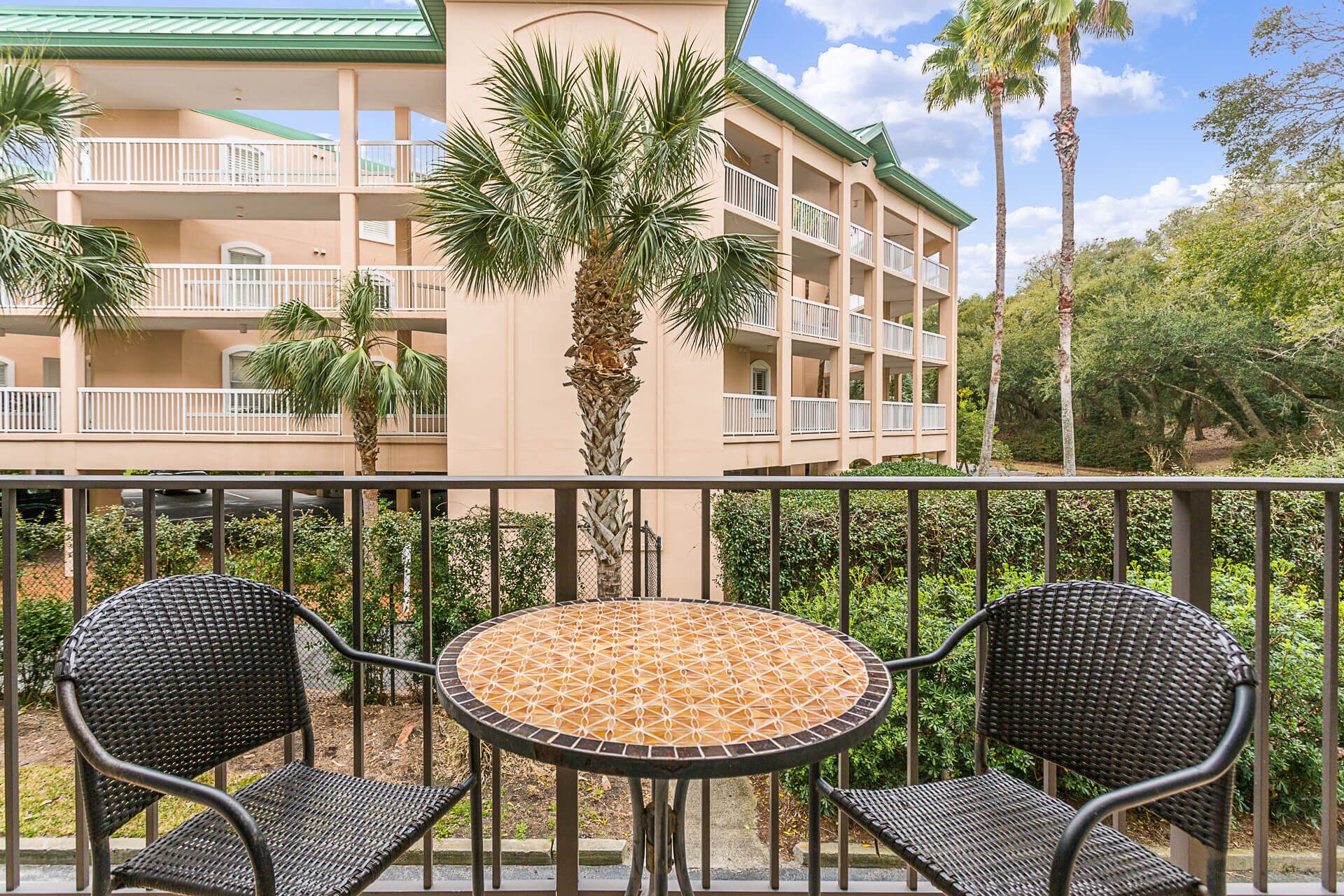 A balcony with two wicker chairs and a mosaic-tiled table, overlooking a building with multiple balconies and palm trees.