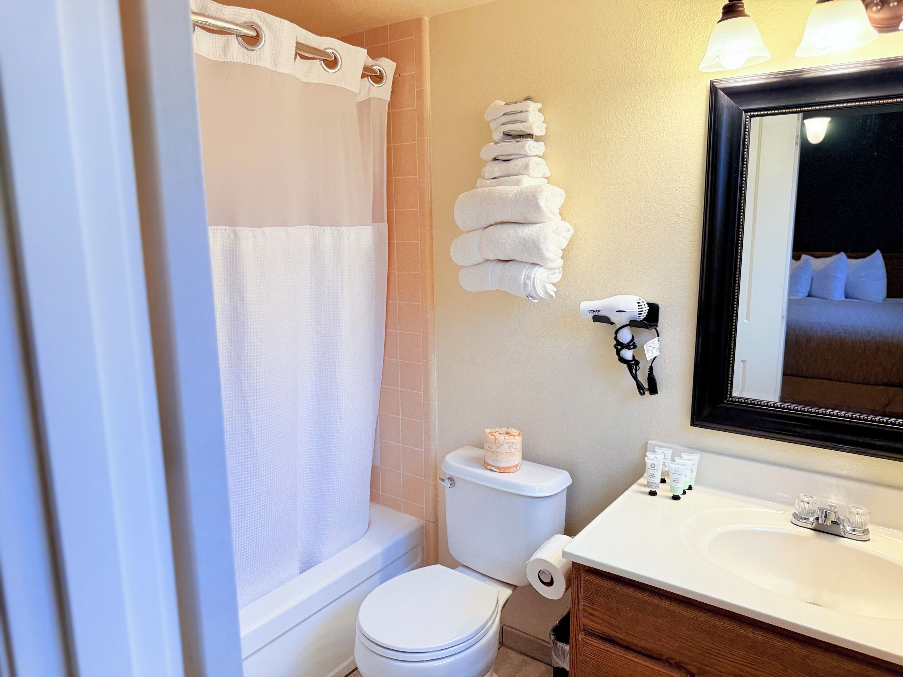 A bathroom with a toilet, a sink, a shower curtain, a stack of towels, a hairdryer, and a mirror.