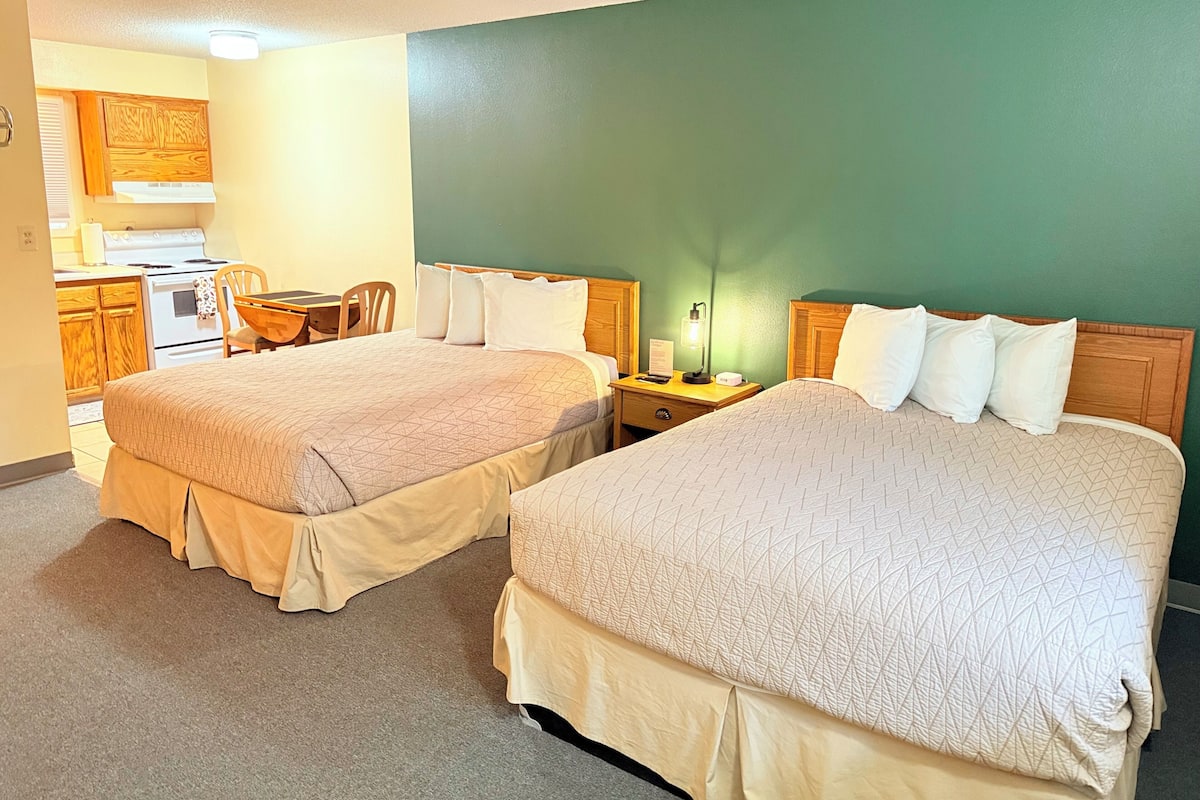 A hotel room with two beds, a desk, a chair, a small kitchenette, and a green wall.