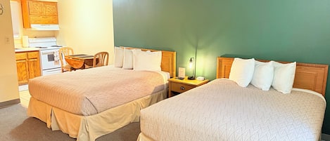 A hotel room with two beds, a desk, a chair, a small kitchenette, and a green wall.