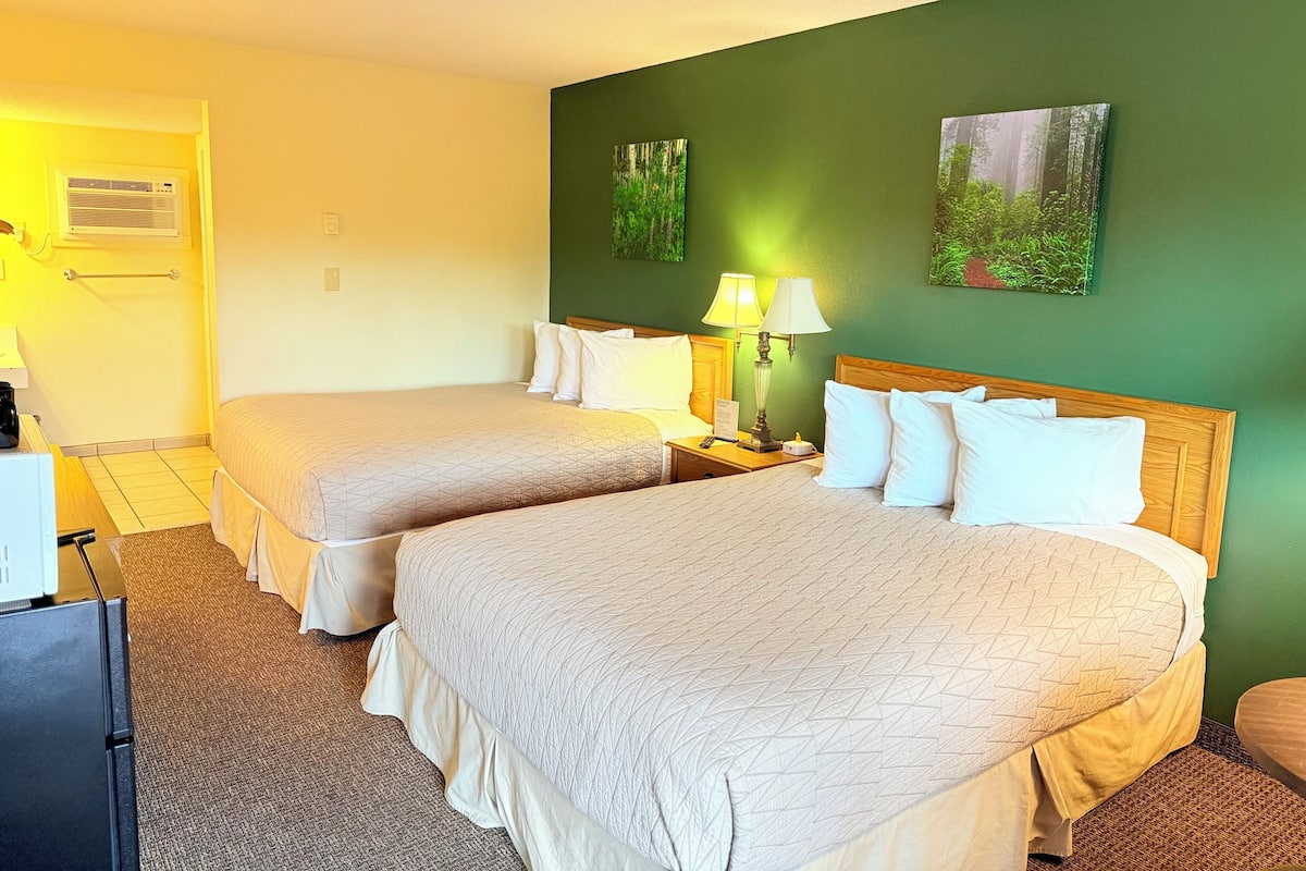 A hotel room with two beds, a nightstand with a lamp, a small table with a coffee maker, and a wall-mounted air conditioner.