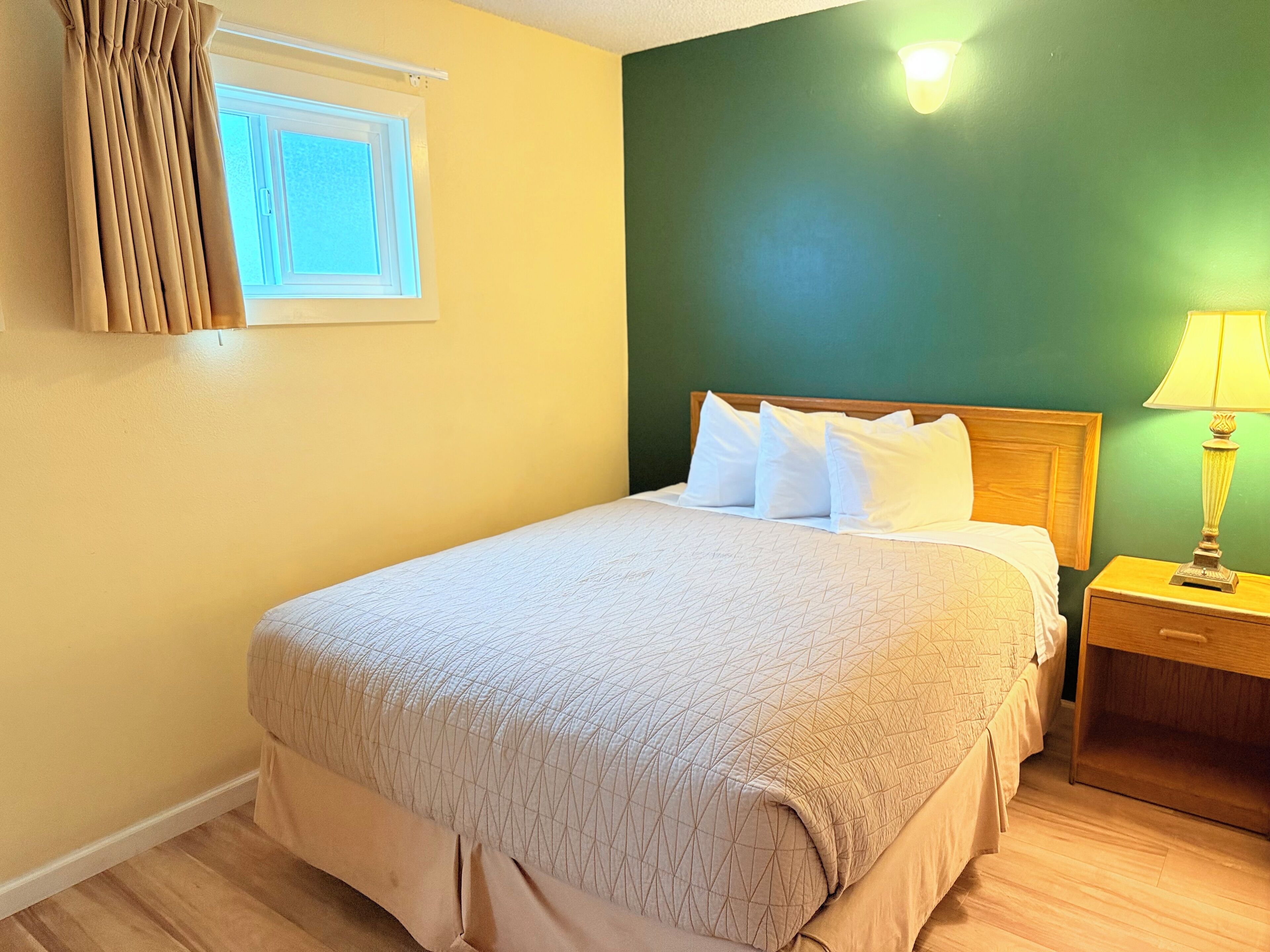 Un lit bien fait, recouvert d’une couvre-lit beige et agrémenté d’oreillers blancs, une table de chevet en bois avec une lampe, et une fenêtre avec des rideaux.