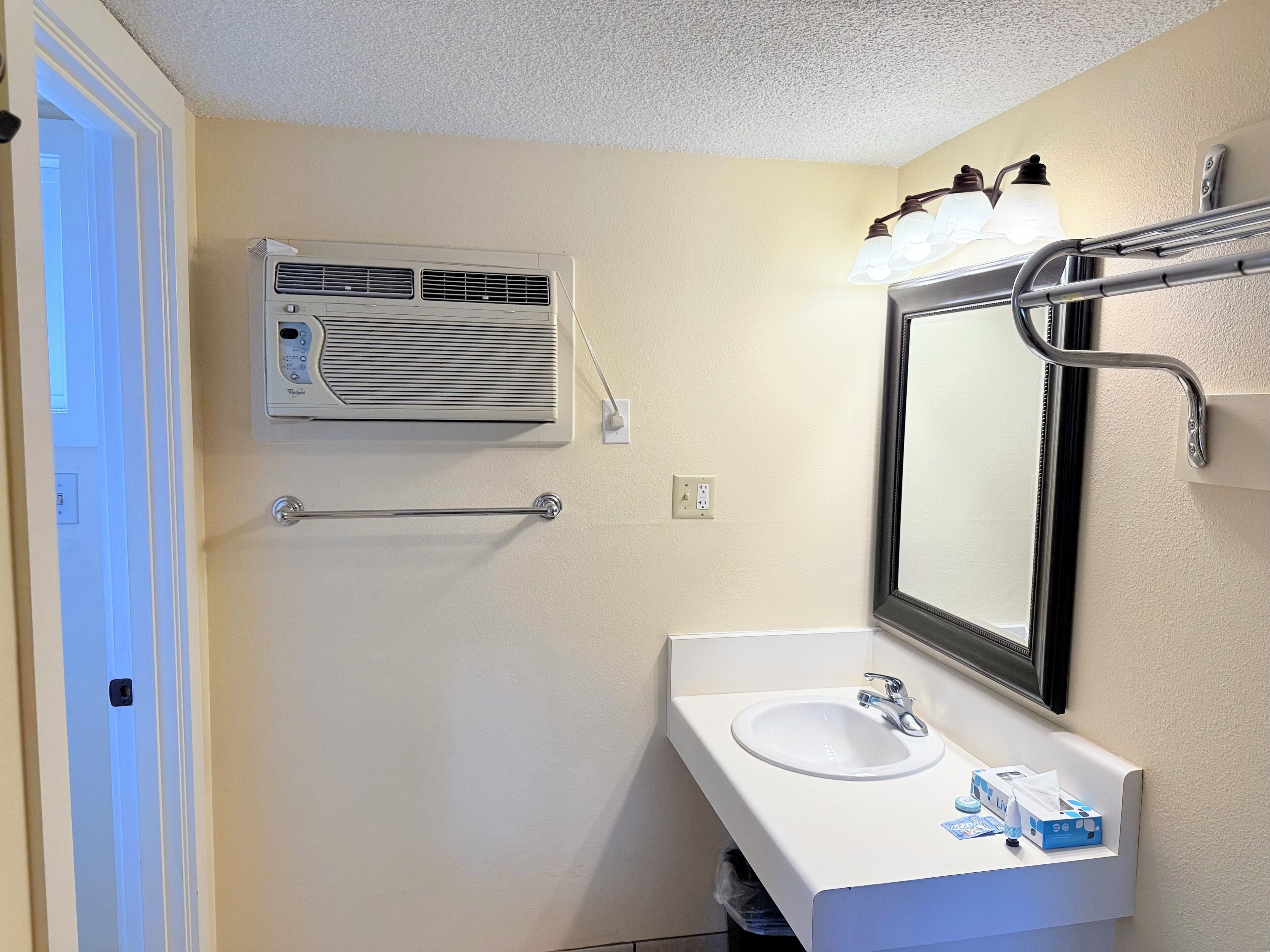 Um banheiro com pia branca, espelho e porta-toalhas. Há um aparelho de ar condicionado fixado na parede e um aparelho de iluminação sobre o espelho.