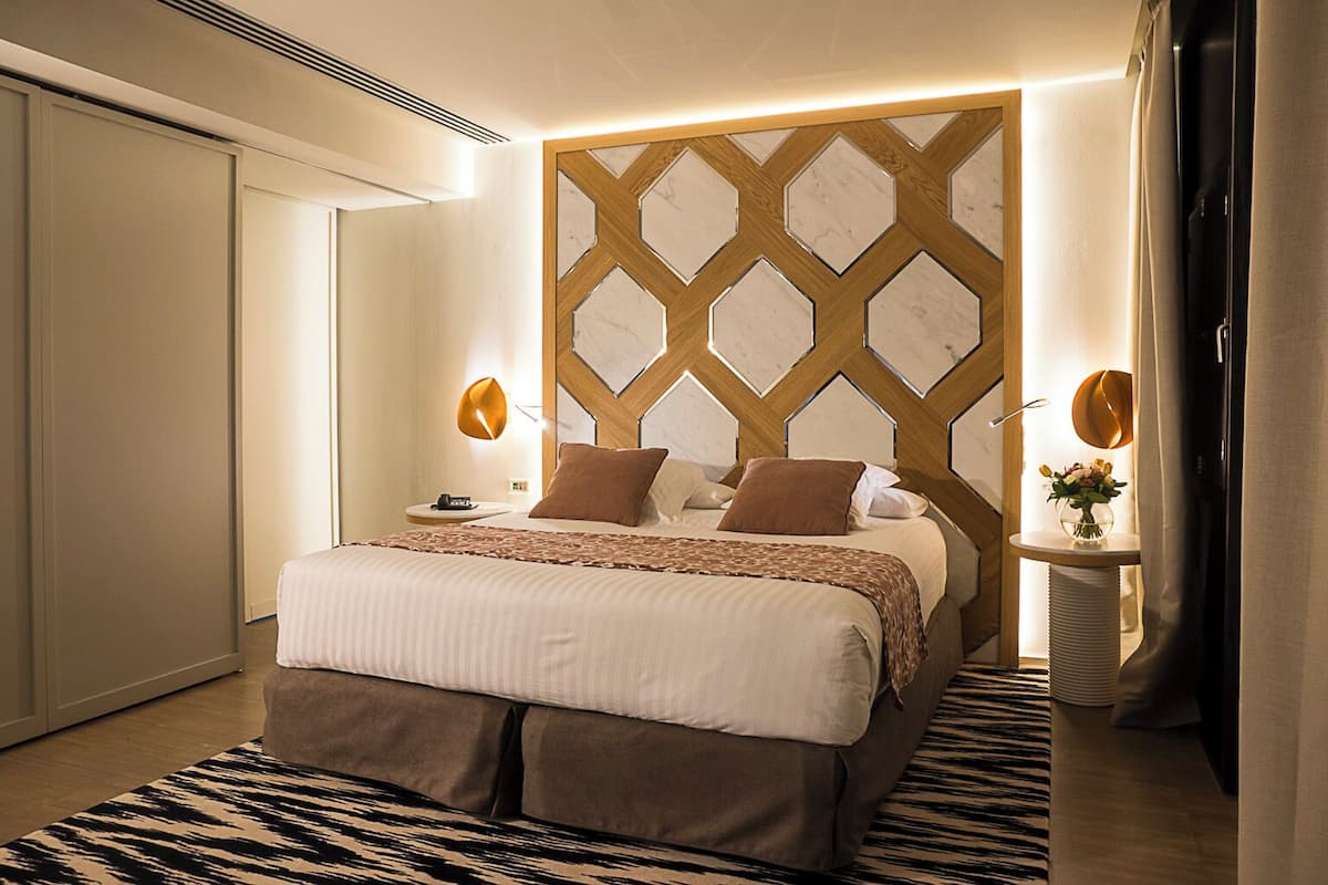 Ein modernes Schlafzimmer mit einem großen Bett, einem geometrischen Wanddesign und einem kleinen Nachttisch mit Blumen in einer Vase.