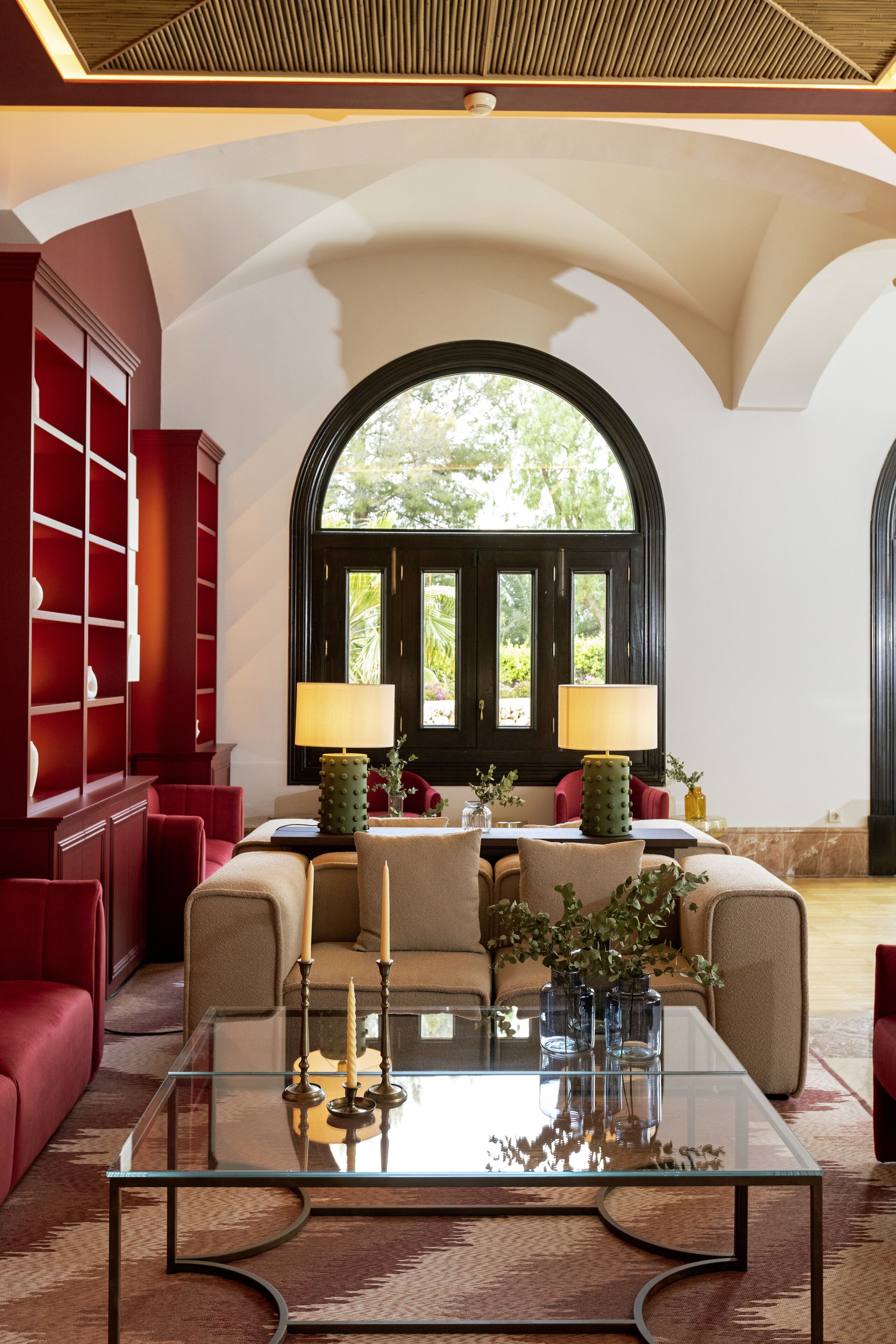 Amplia sala de estar con una mesa de centro de vidrio, muebles rojos y un gran ventanal de arco que ofrece vistas al verdor exterior.