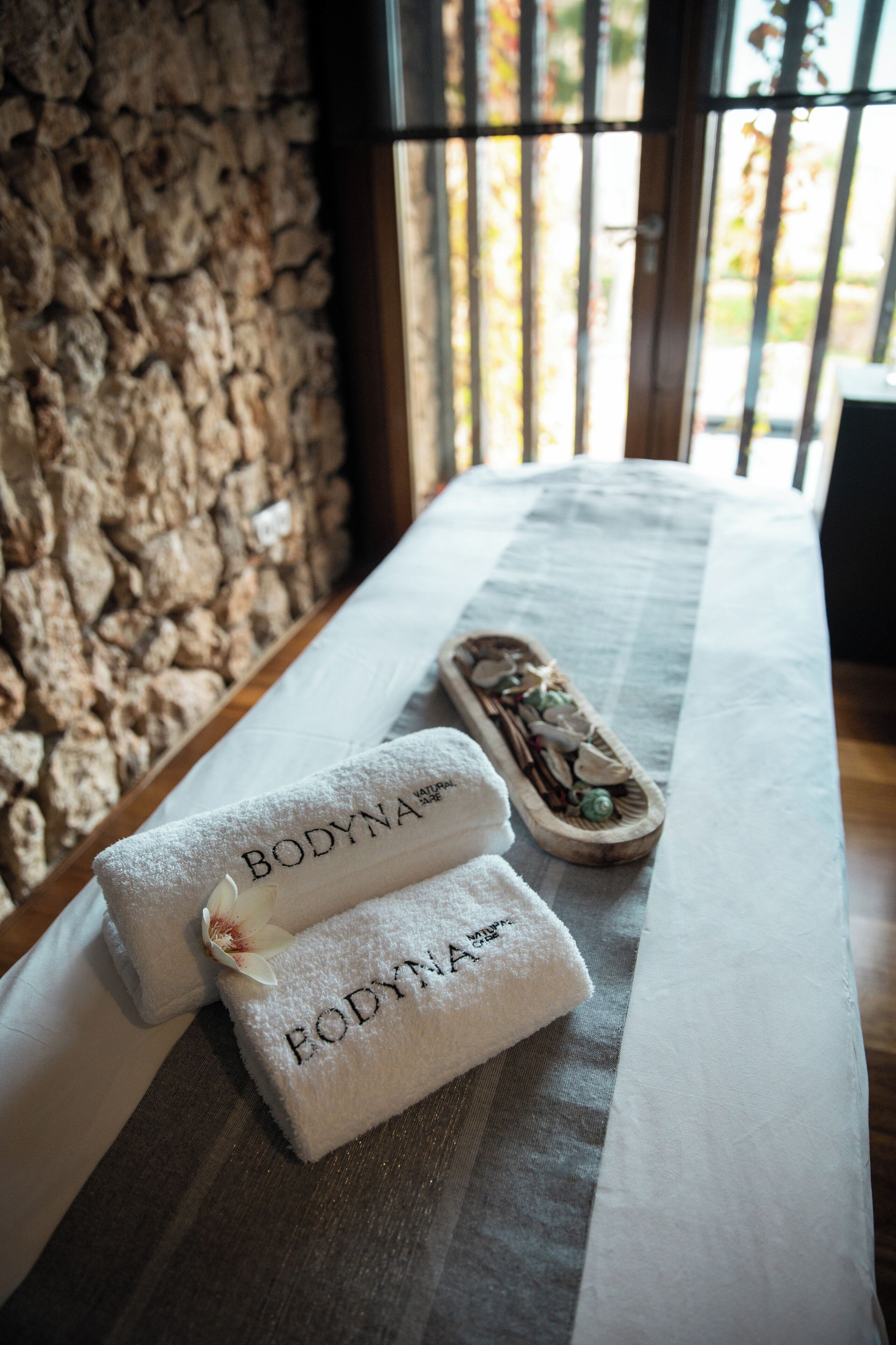 Una cama bien hecha con dos toallas blancas con el rótulo "BODYNA" y una bandeja de madera con piedras decorativas.