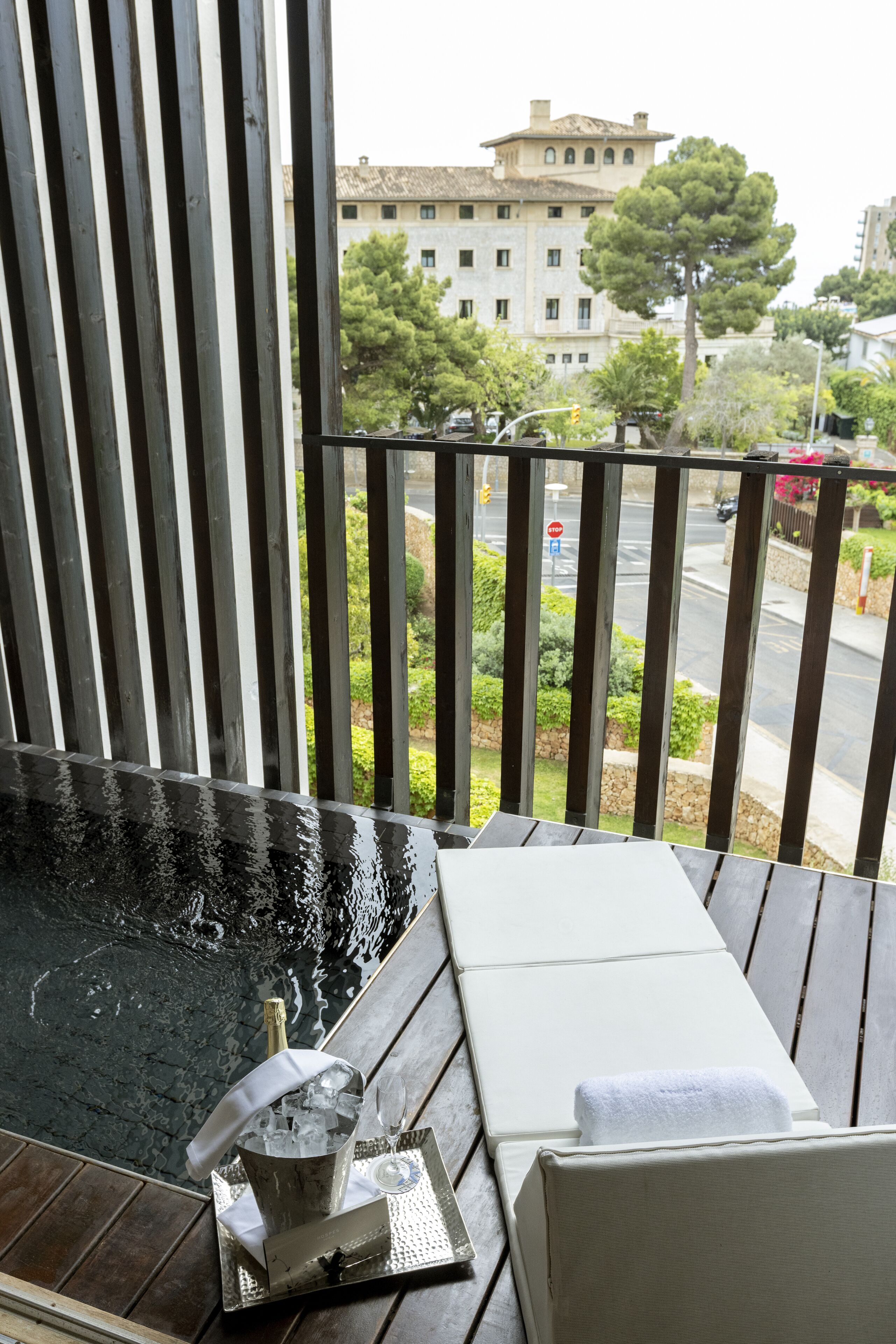 Un balcon avec un jacuzzi, un plateau avec une bouteille et des verres, et une vue sur une rue et des immeubles.