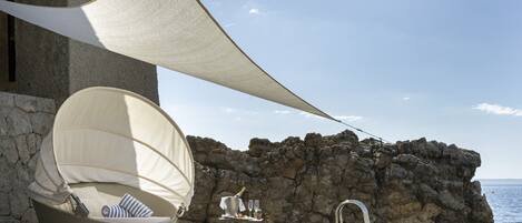 A seaside patio with a wicker chair, a table with glasses and a bottle, and a rocky backdrop.