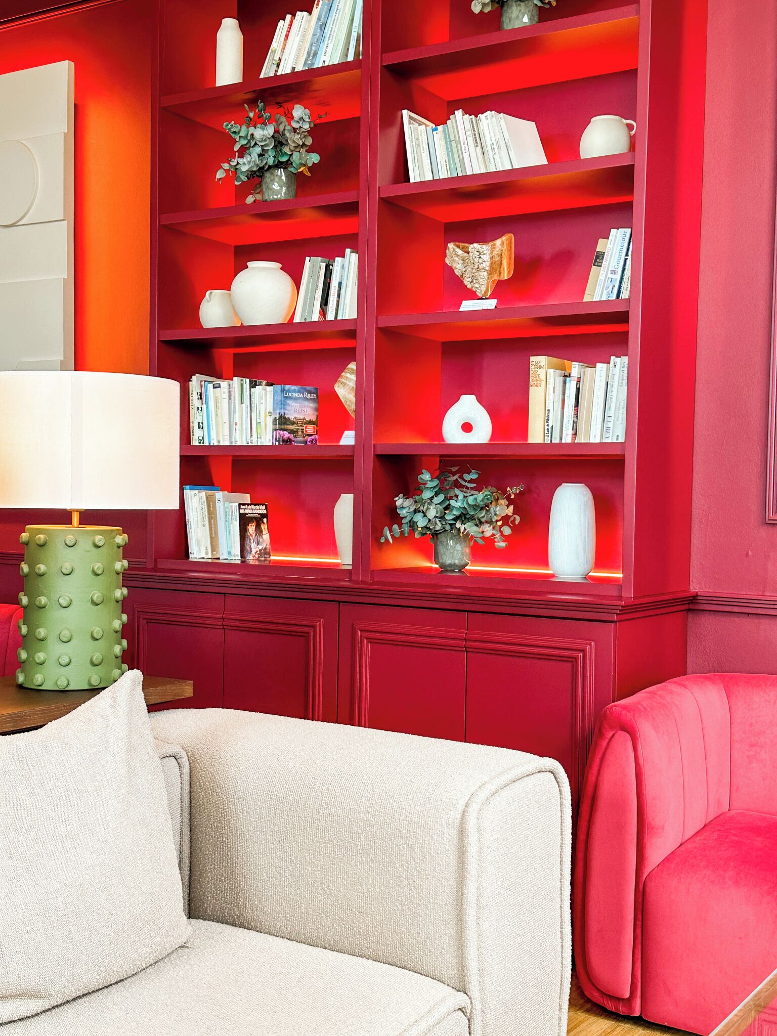 A living room with a red bookshelf filled with books and decorative items, a beige sofa, and a green lamp.