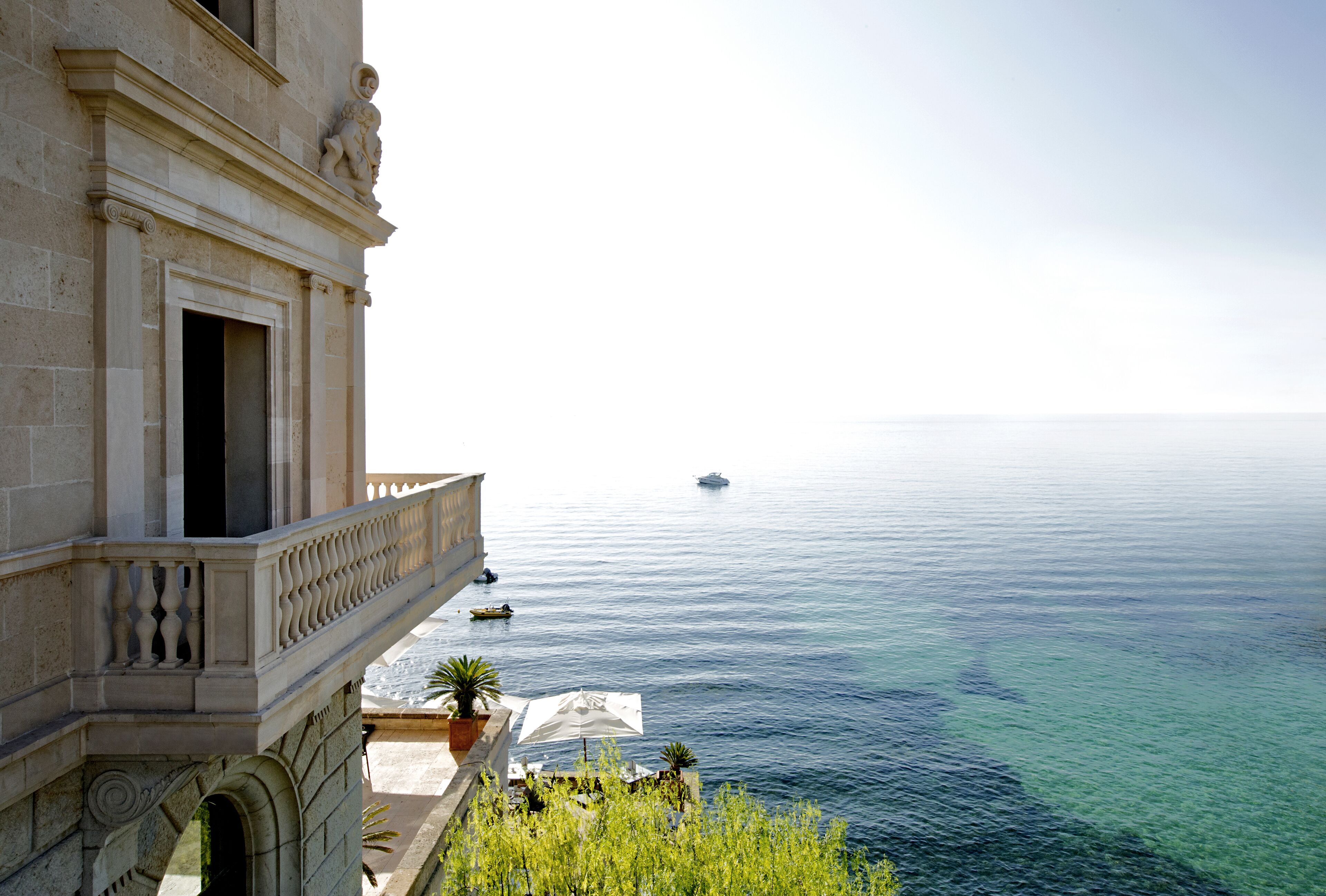 A building with a balcony overlooking a calm sea, a small boat in the distance, and a clear sky.