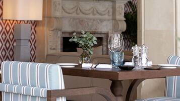 A dining area with striped upholstered chairs, a table set with plates, glasses, and a vase of flowers. A fireplace with a decorative mantle is in the background.
