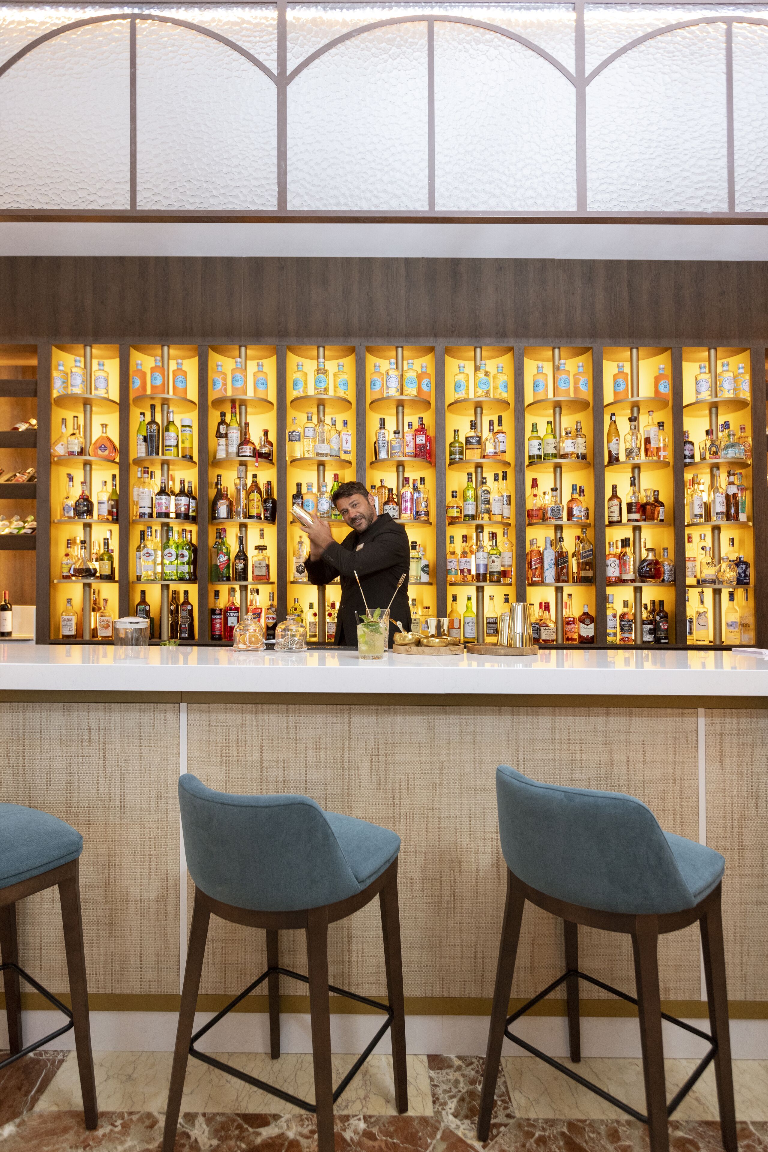 A bar area with shelves of liquor bottles, a bartender preparing drinks, and blue bar stools.