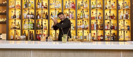 A bar area with shelves of liquor bottles, a bartender preparing drinks, and blue bar stools.