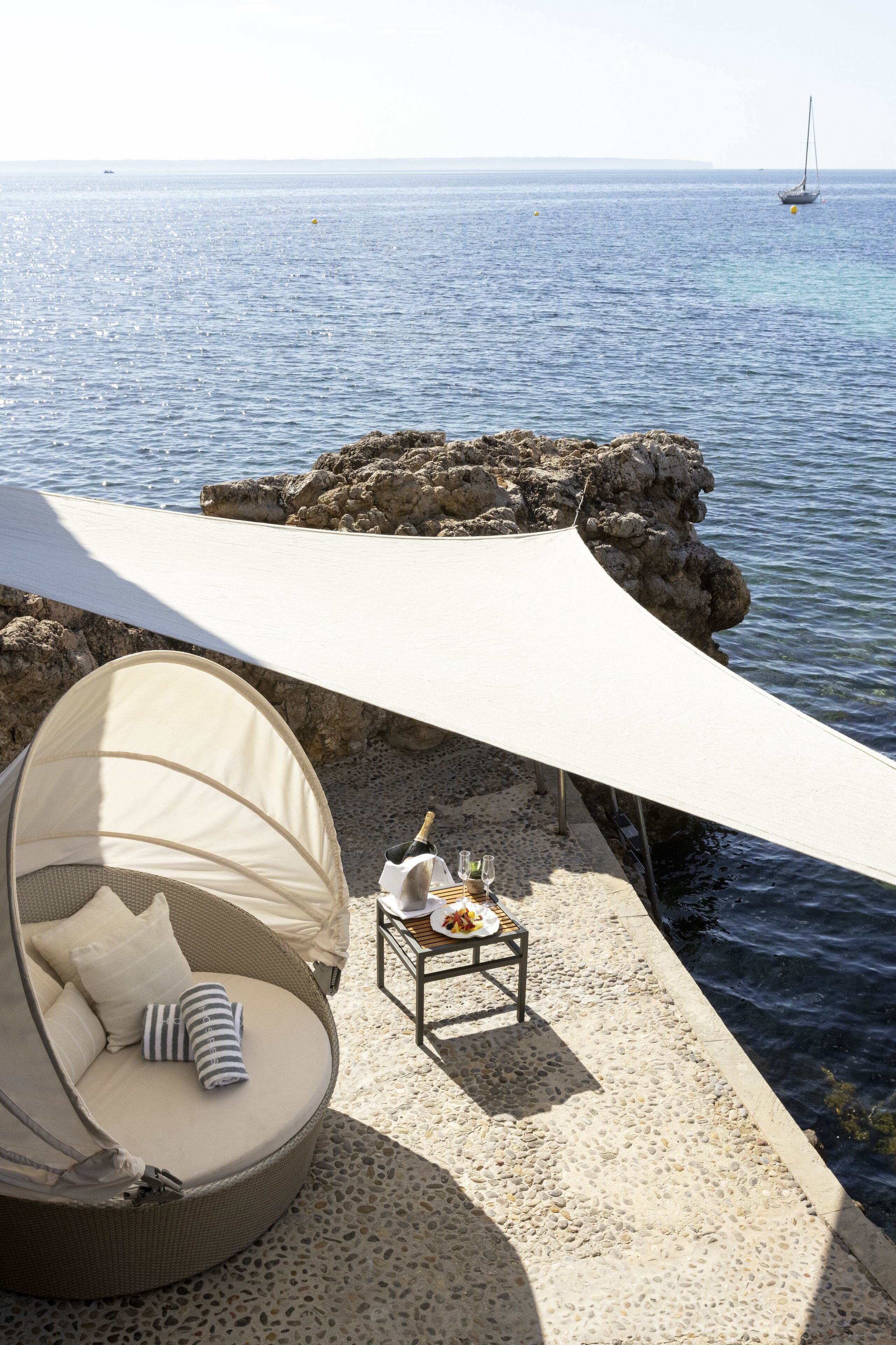 A beachside seating area with a cushioned chair and a table set with a bottle and glasses, overlooking the sea.
