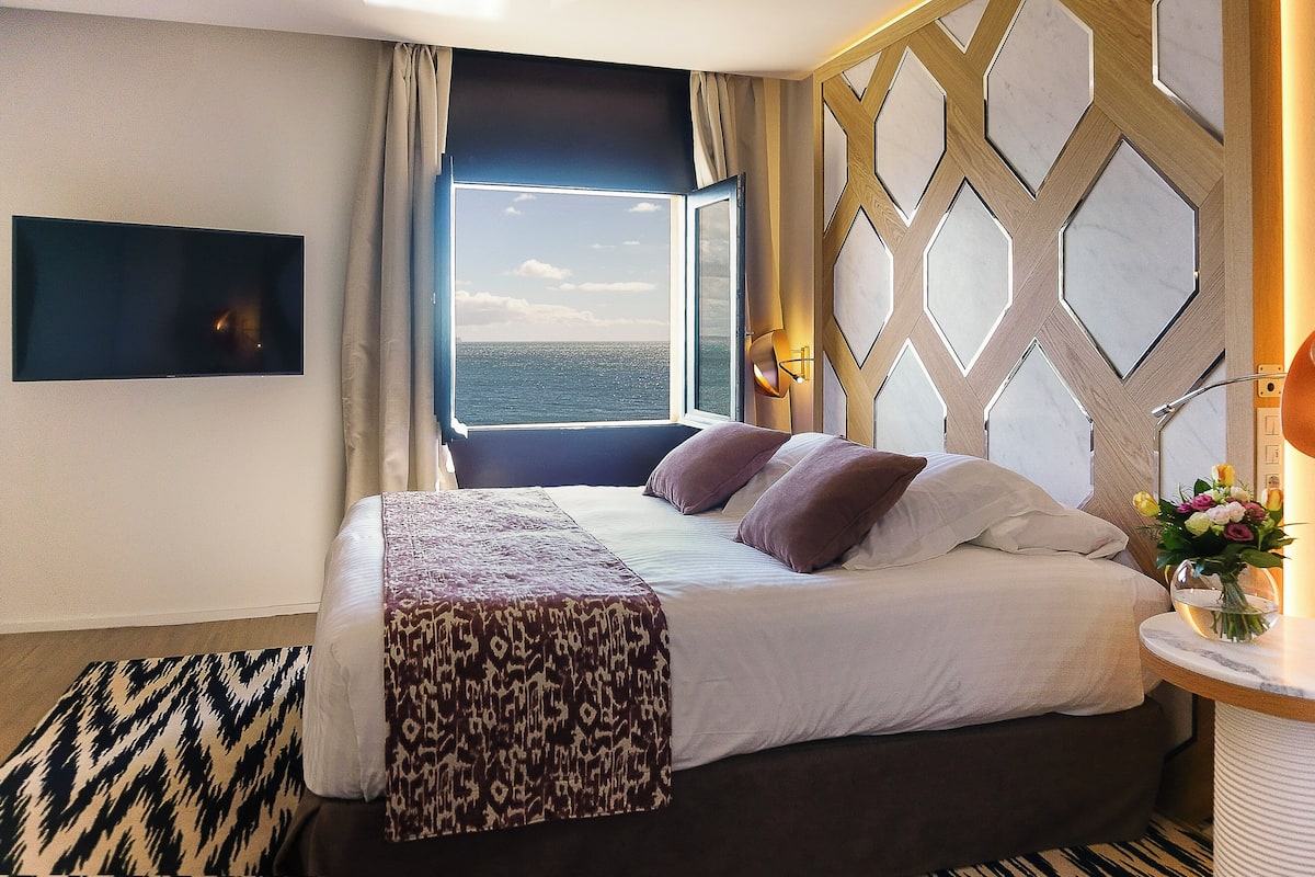 Ein Hotelzimmer mit einem großen Bett, Meerblick durch ein Fenster, einem wandmontierten Flachbildfernseher und einer dekorativen Wandverkleidung.