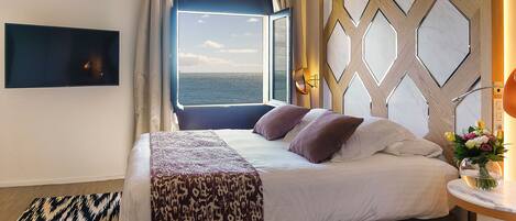 A hotel room with a large bed, a view of the sea through a window, a flat-screen TV mounted on the wall, and a decorative wall panel.