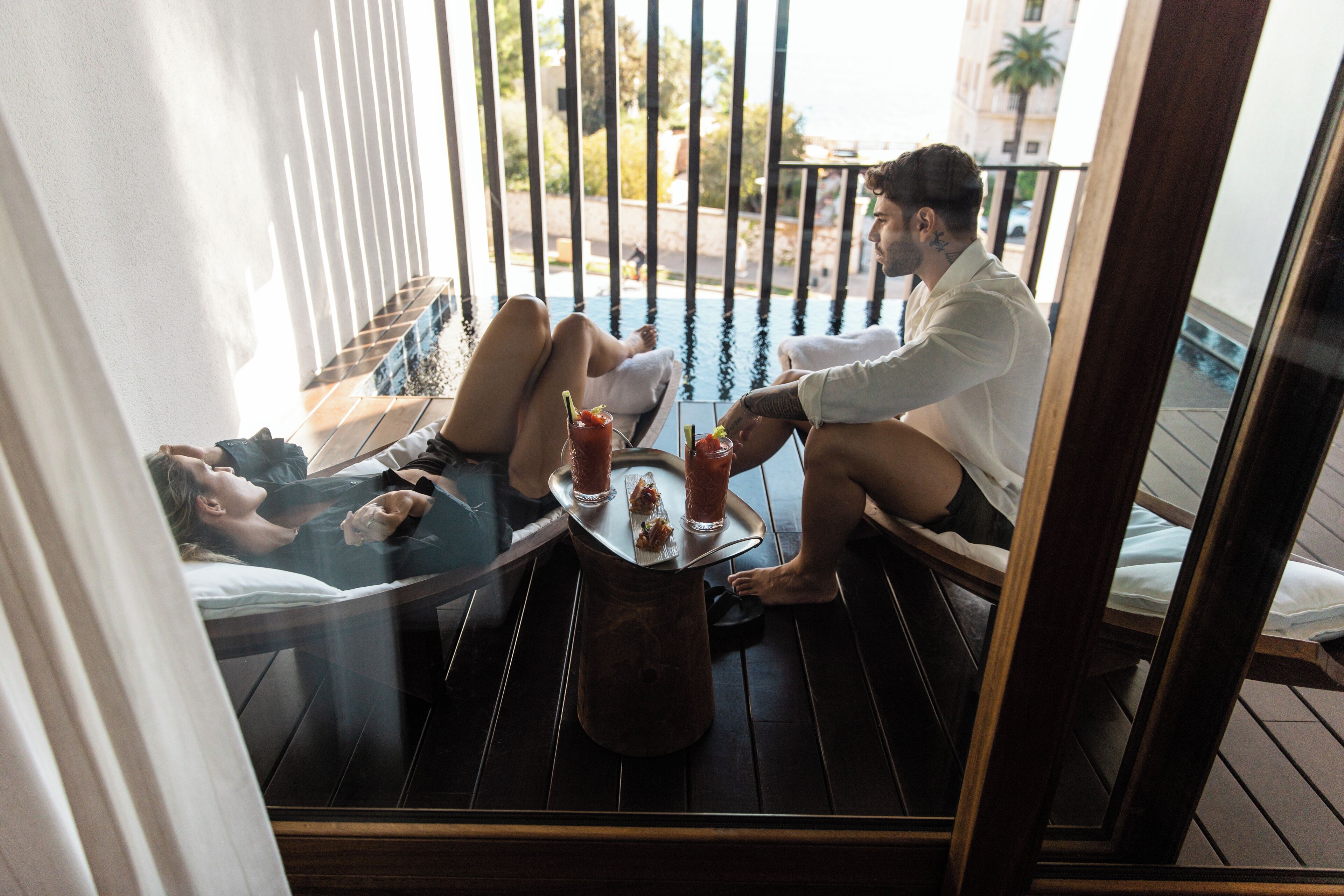 Een koppel ontspant op een balkon met een glazen tafel, een stoel en uitzicht op de straat en gebouwen.