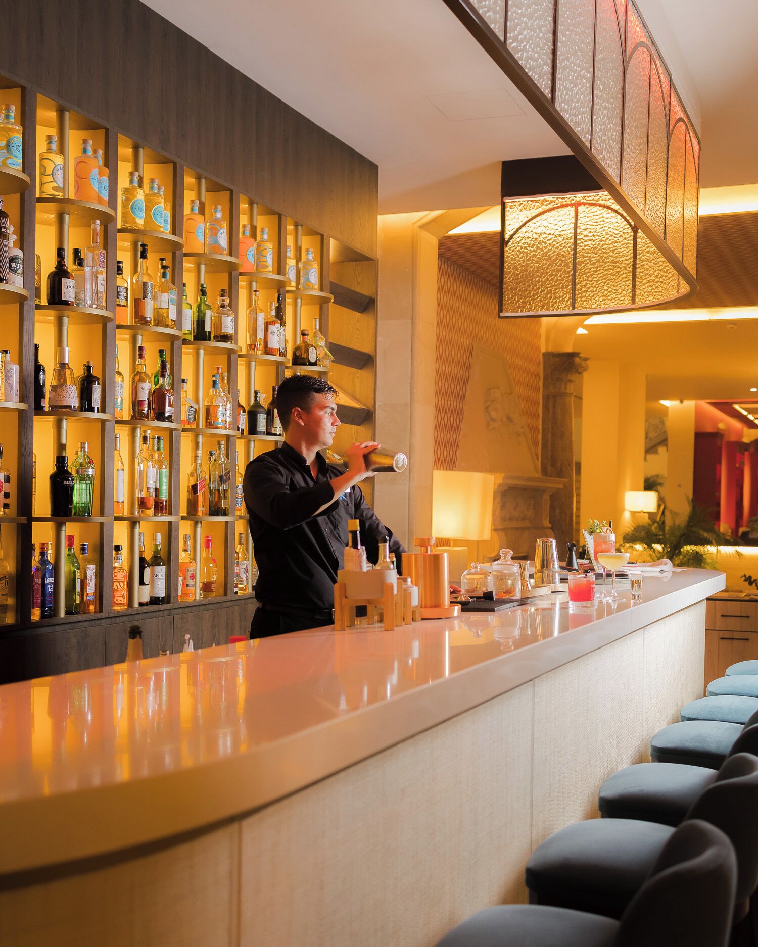 Zona de bar con estantes de botellas de licor, un bartender preparando una bebida y asientos de diseño moderno.