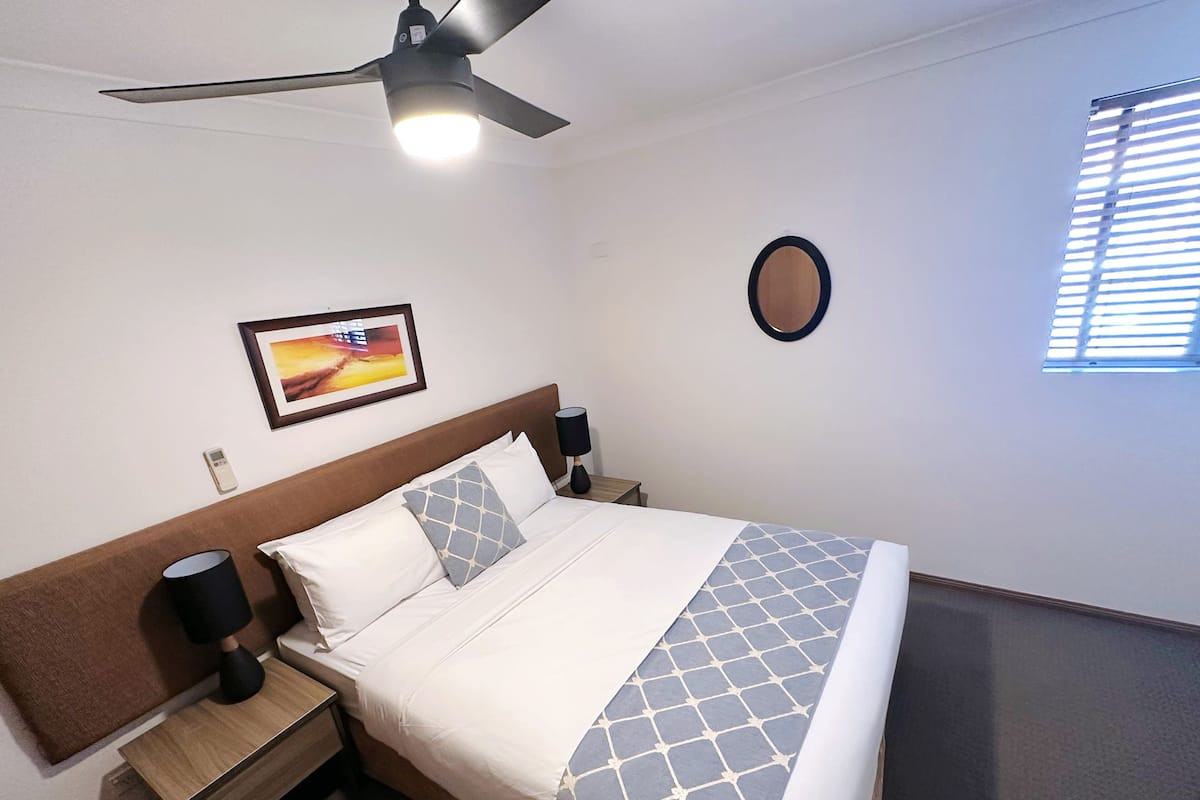Ein ordentlich bezogenes Bett mit einem gemusterten Überwurf, ein Deckenventilator, eine Nachttischlampe, ein gerahmtes Bild an der Wand und ein kleiner Tisch neben dem Bett.