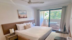 Habitación de hotel con cama, mesita de noche, escritorio, silla, ventilador de techo, un cuadro y vistas al exterior a través de una puerta de vidrio.