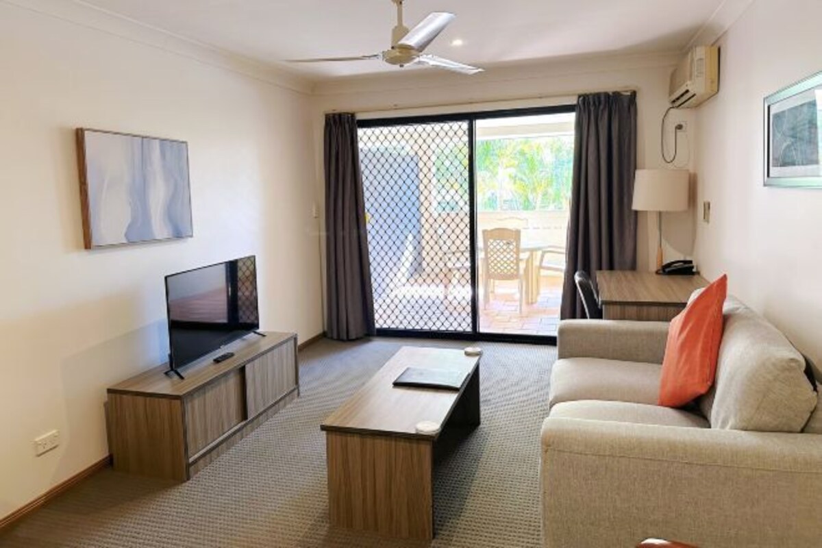 A modern hotel room with a flat-screen TV, a wooden coffee table, a sofa, and a sliding glass door leading to an outdoor area.