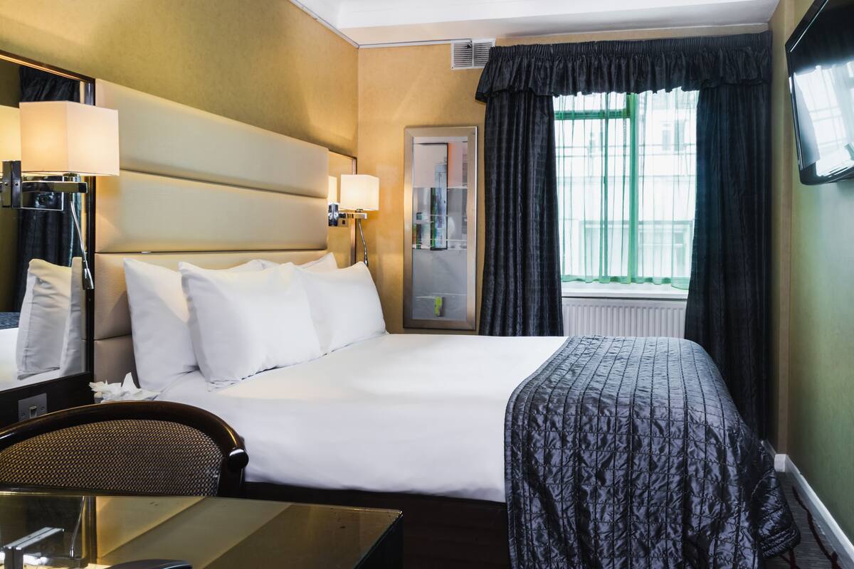 Une chambre d’hôtel avec un grand lit, un bureau avec une chaise, un miroir, une fenêtre avec des rideaux et un téléviseur fixé au mur.