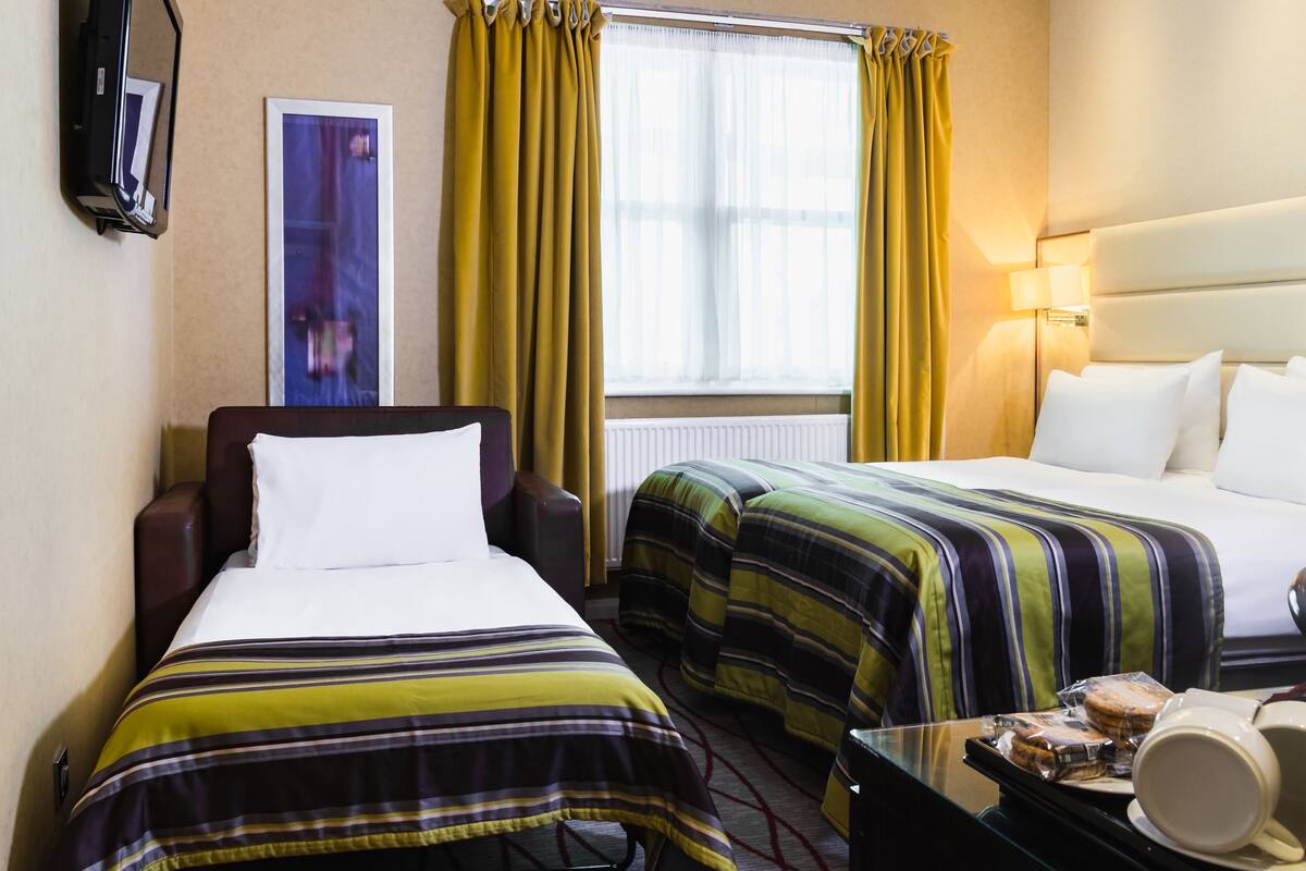 Une chambre d’hôtel avec deux lits, une télévision, une fenêtre avec des rideaux, une lampe et une petite table sur laquelle se trouvent une tasse et une assiette.